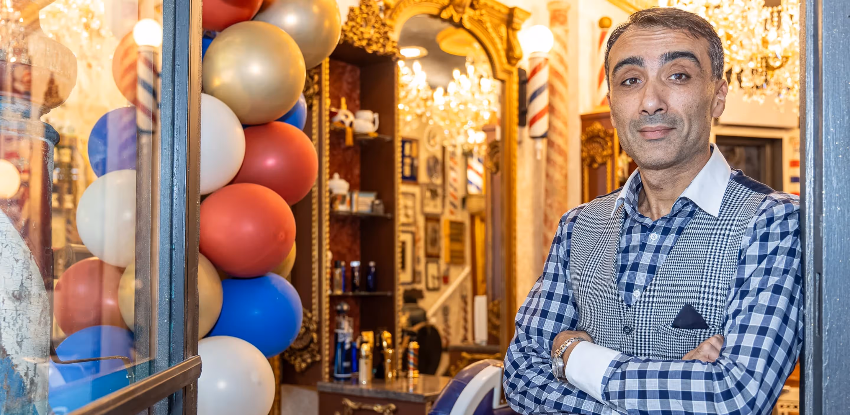 Barbershop owner Artur standing at the entrance of a vintage-style barbershop with festive red, white, and blue balloons and ornate interior decor
