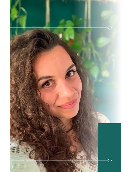 Portrait einer Frau mit lockigen Haaren die symphatisch in die Kamera lächelt. Im Hintergrund sieht man eine grüne Wand mit Hängepflanzen.