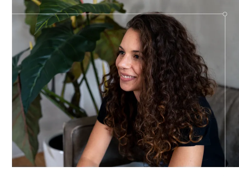 Porträt einer Frau mit lockigem Haar, die entspannt auf einem Sofa sitzt und freundlich lächelt. Im Hintergrund sorgt eine große Zimmerpflanze für eine natürliche, warme Atmosphäre.