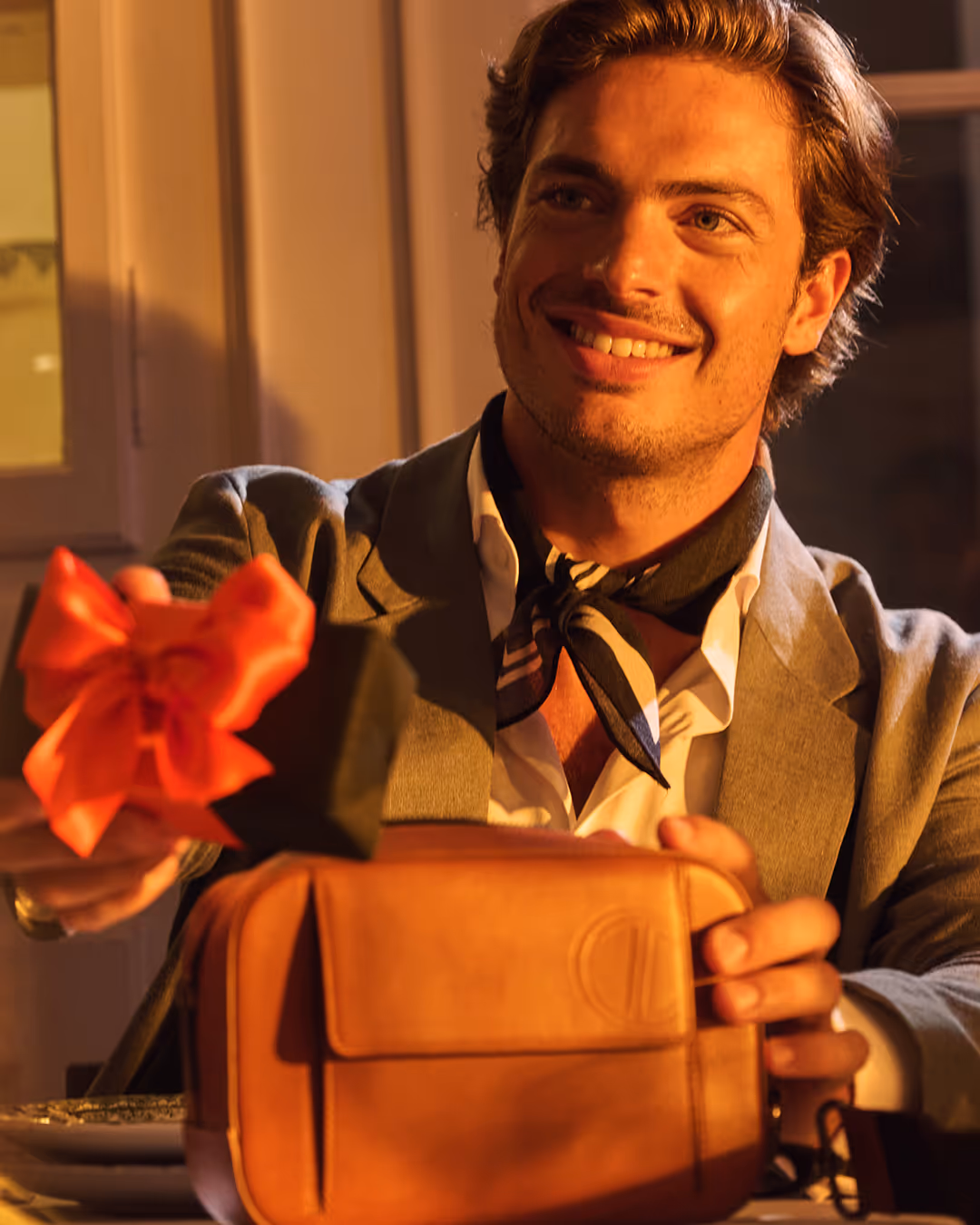 Smiling man in a blazer and scarf holding a small black gift box with a red bow and a brown leather bag.