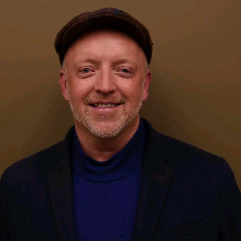 Smiling man with light beard wearing a dark flat cap, dark blazer, and blue turtleneck sweater against a brown background.