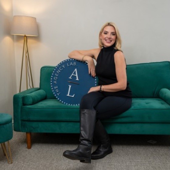 Smiling woman with blonde hair in black outfit and boots sitting on a green velvet couch, holding a blue round sign that reads 'The Agency Lab'.