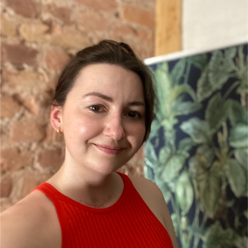 Smiling woman with dark hair wearing a red sleeveless top, standing indoors with a brick wall and leafy patterned wallpaper in the background.