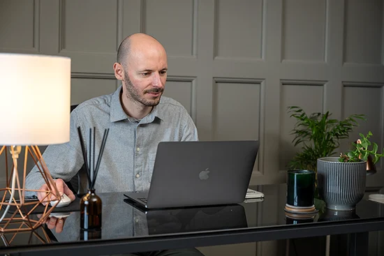 Man working on a laptop at a black desk with a lamp and potted plants.