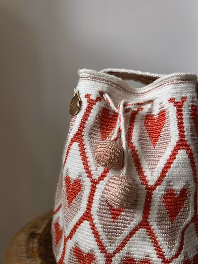 White crocheted bag with red heart patterns and two beige pom-pom tassels hanging from the handle.