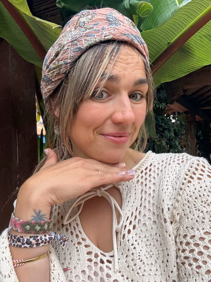 Smiling woman wearing a patterned headscarf and a white crochet top, posing with her hand under her chin in front of large green leaves.