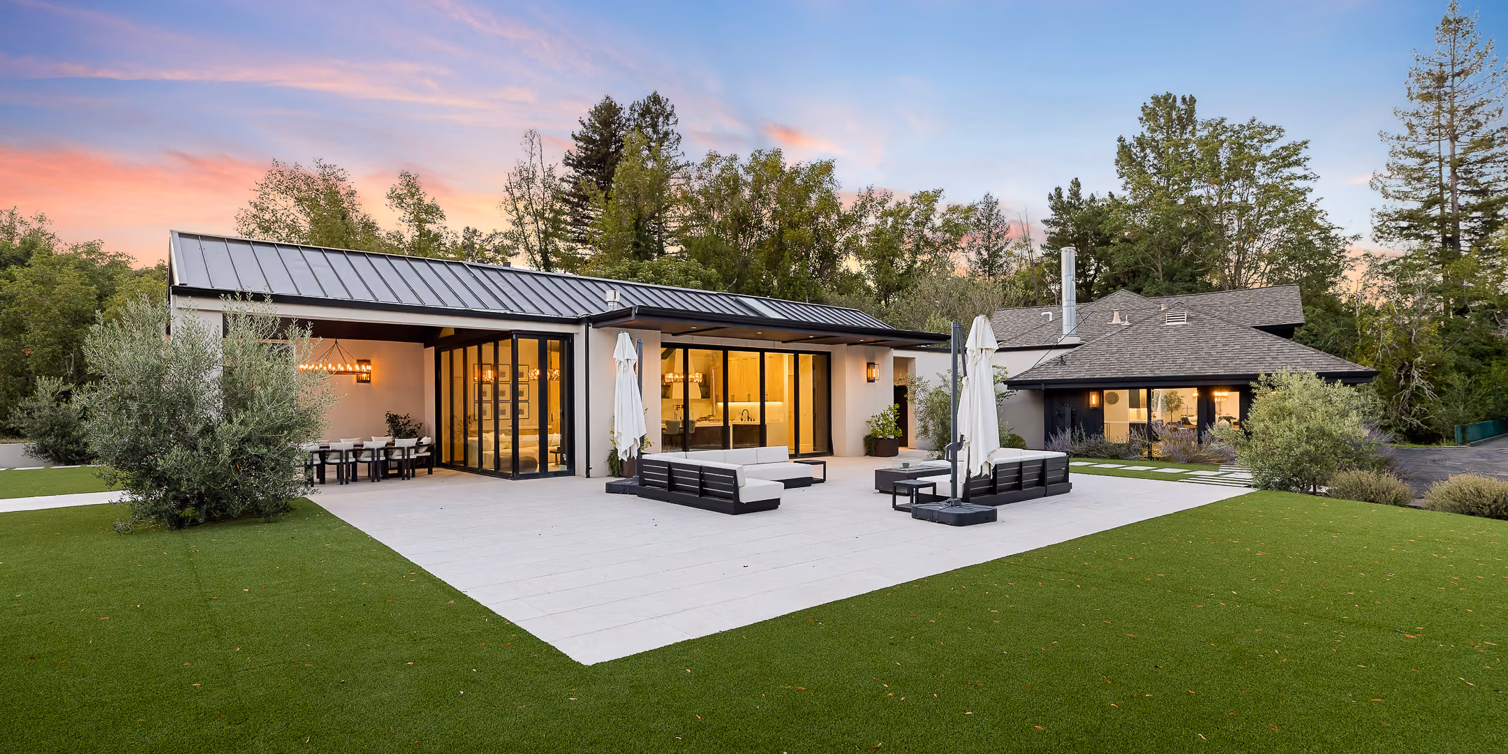 Modern house with large glass doors opening to patio with seating and green lawn at sunset.