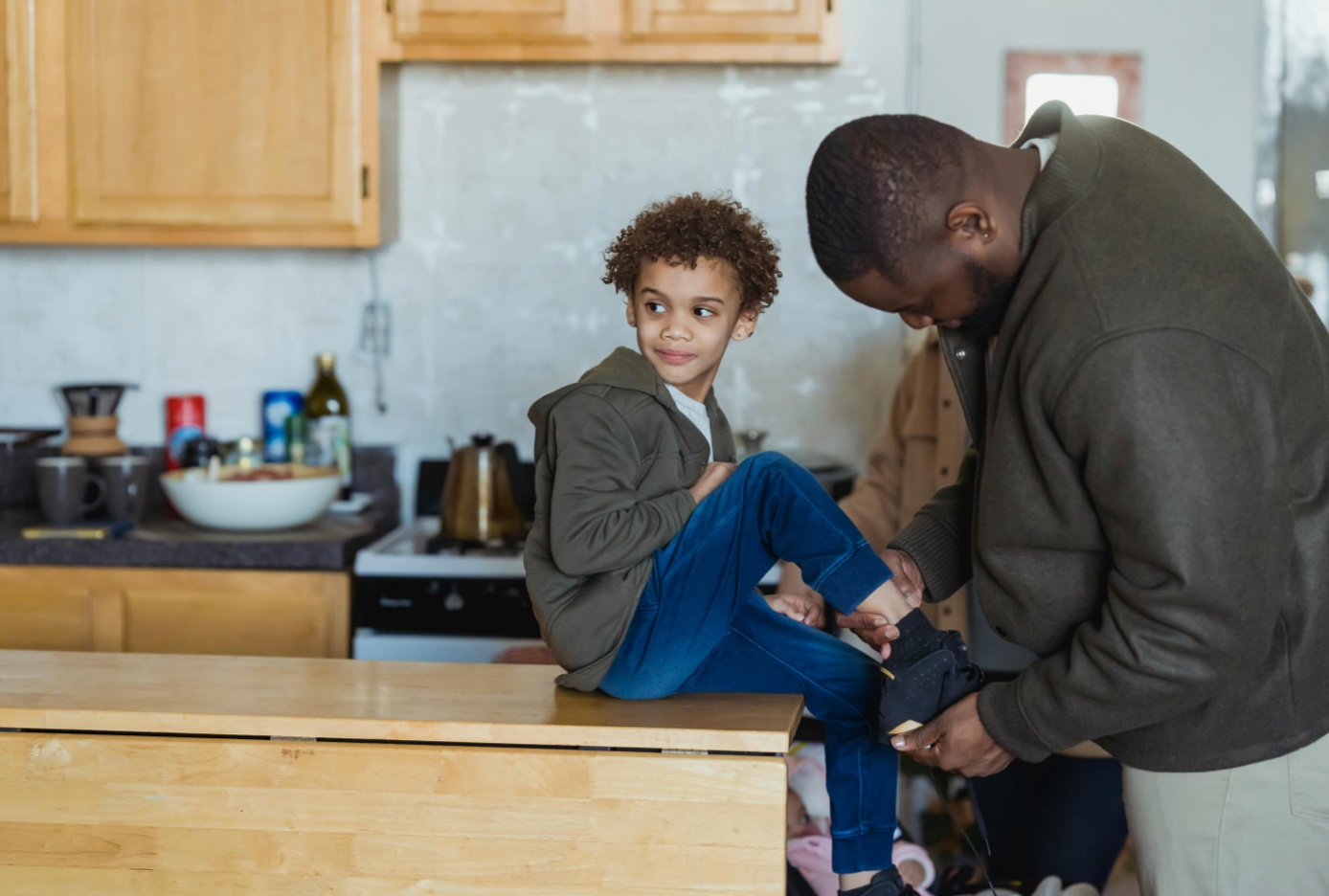 Father tying his son's shoes while the child sits on a kitchen counter.