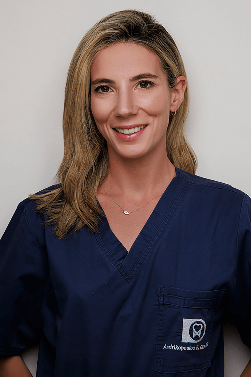 Smiling woman with shoulder-length blonde hair wearing navy blue medical scrubs and a delicate necklace.