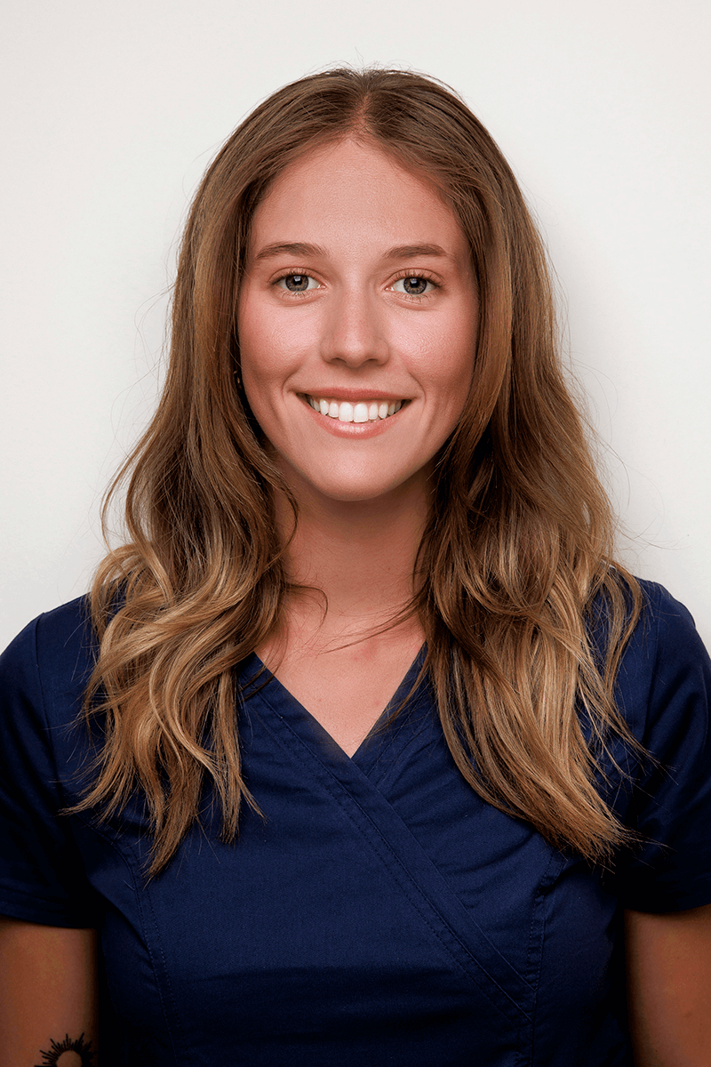 Smiling woman with long wavy brown hair wearing a navy blue scrub top against a plain light background.