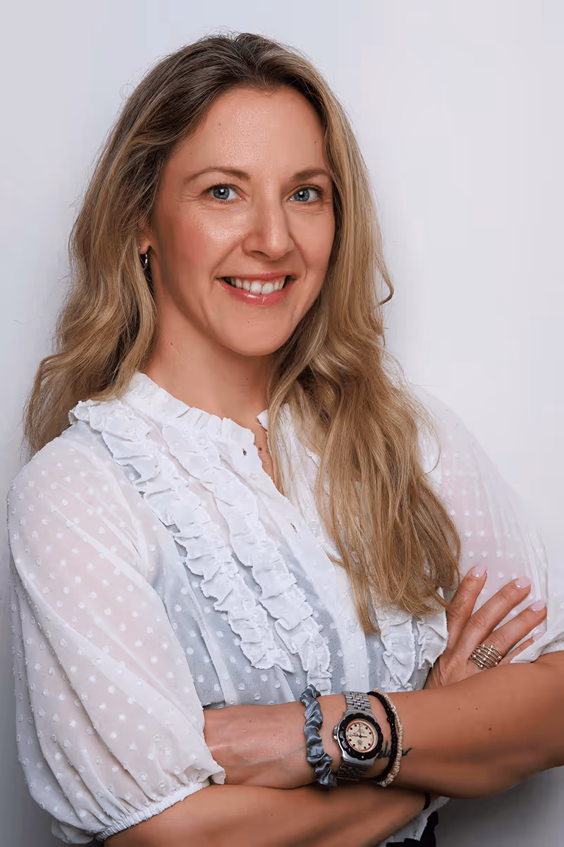 Smiling woman with long blonde hair wearing a white ruffled blouse with arms crossed.