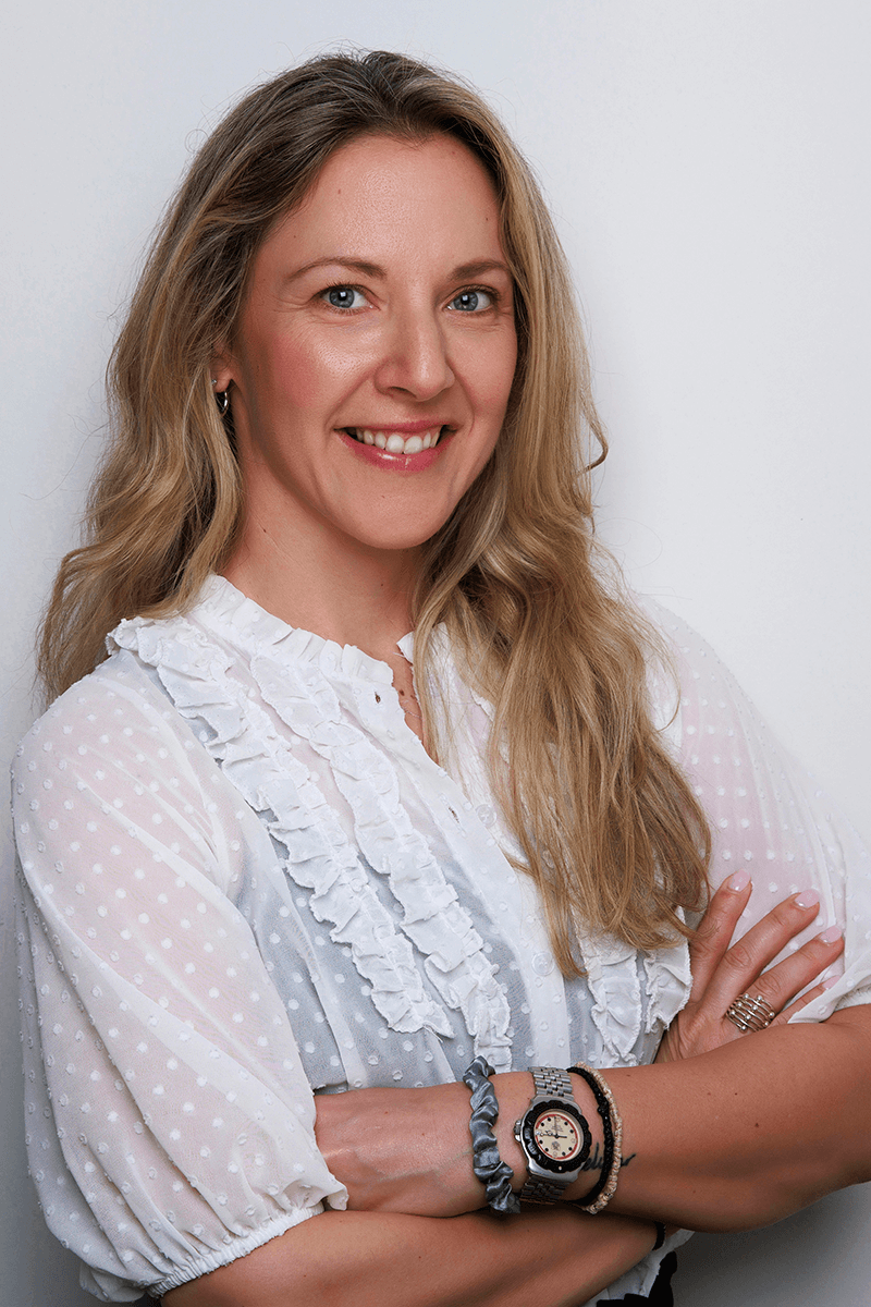 Smiling woman with long blonde hair wearing a white ruffled blouse with arms crossed.