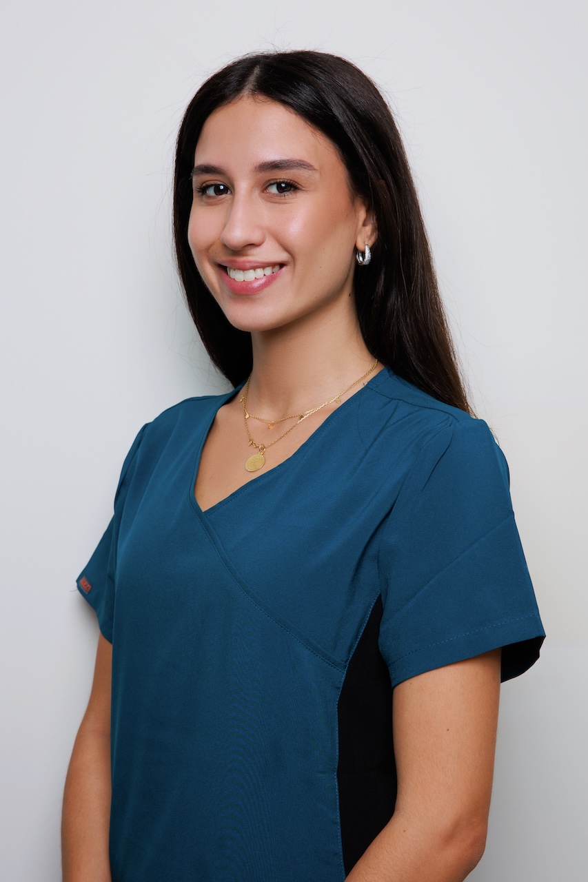 Smiling young woman with long dark hair wearing a teal scrub top and gold necklaces against a plain light background.