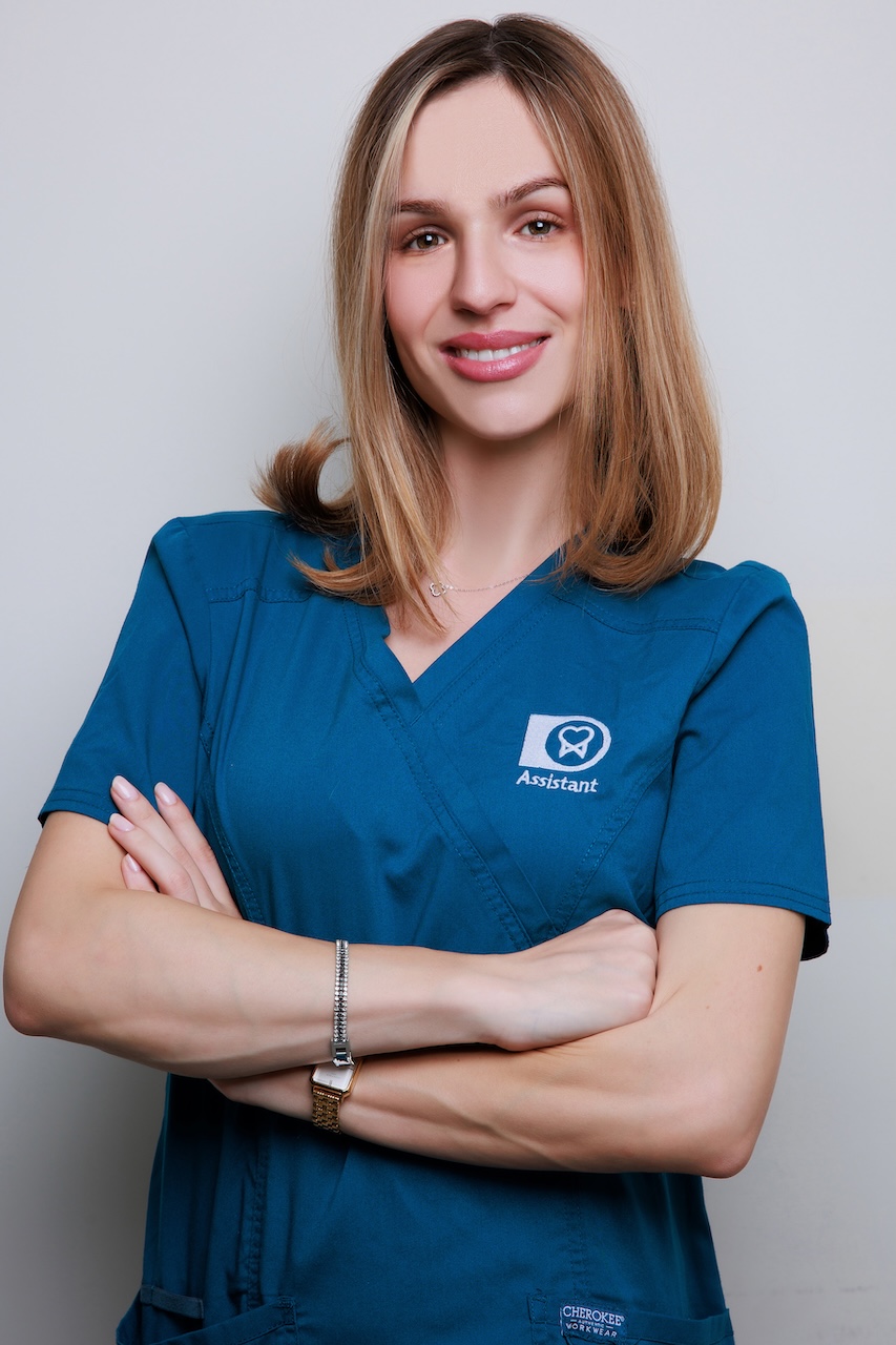 Smiling woman in blue medical scrubs with arms crossed against a light gray background.