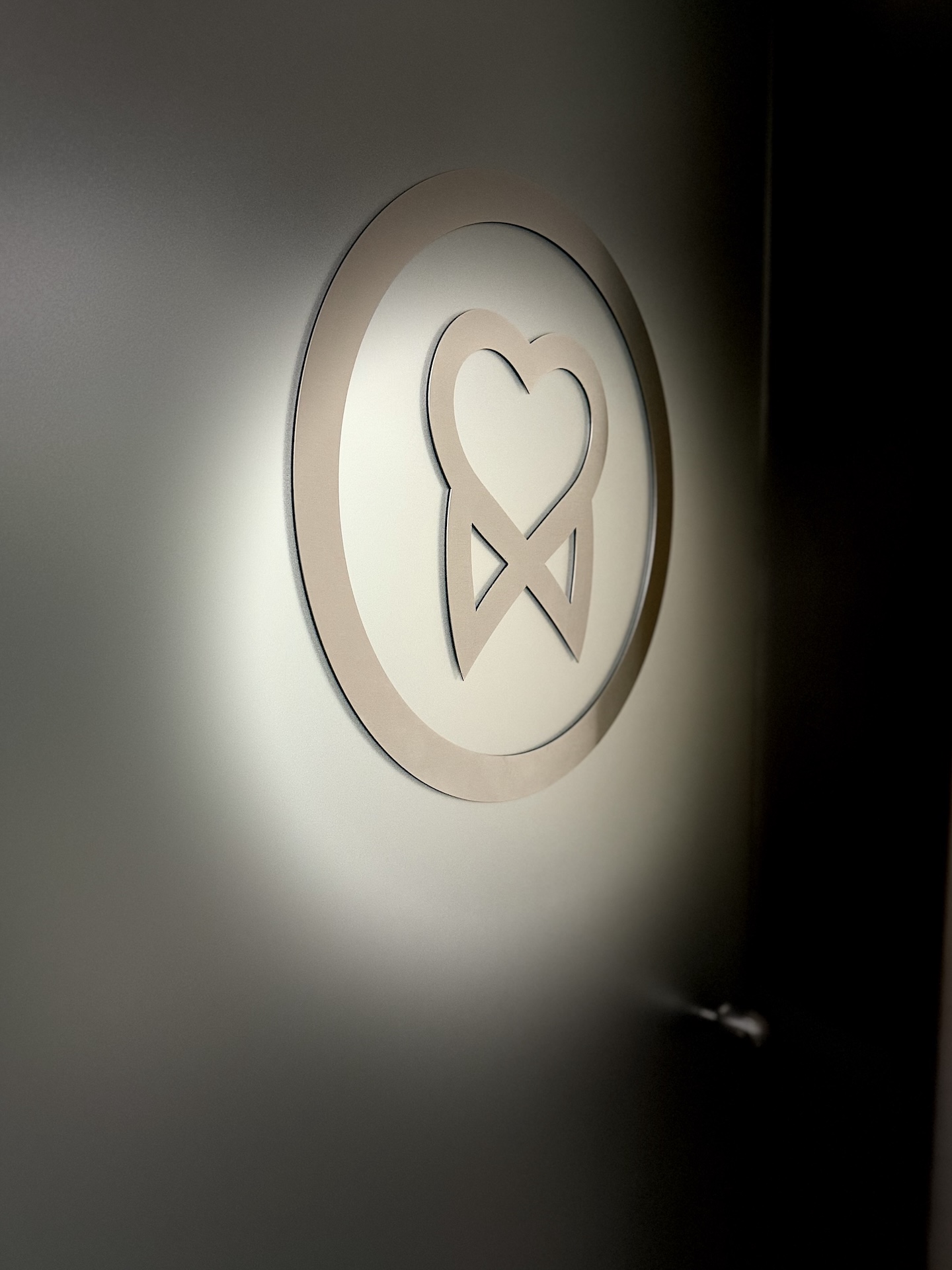 Wall-mounted circular sign with a heart-shaped emblem inside a ring, softly illuminated against a dark background.