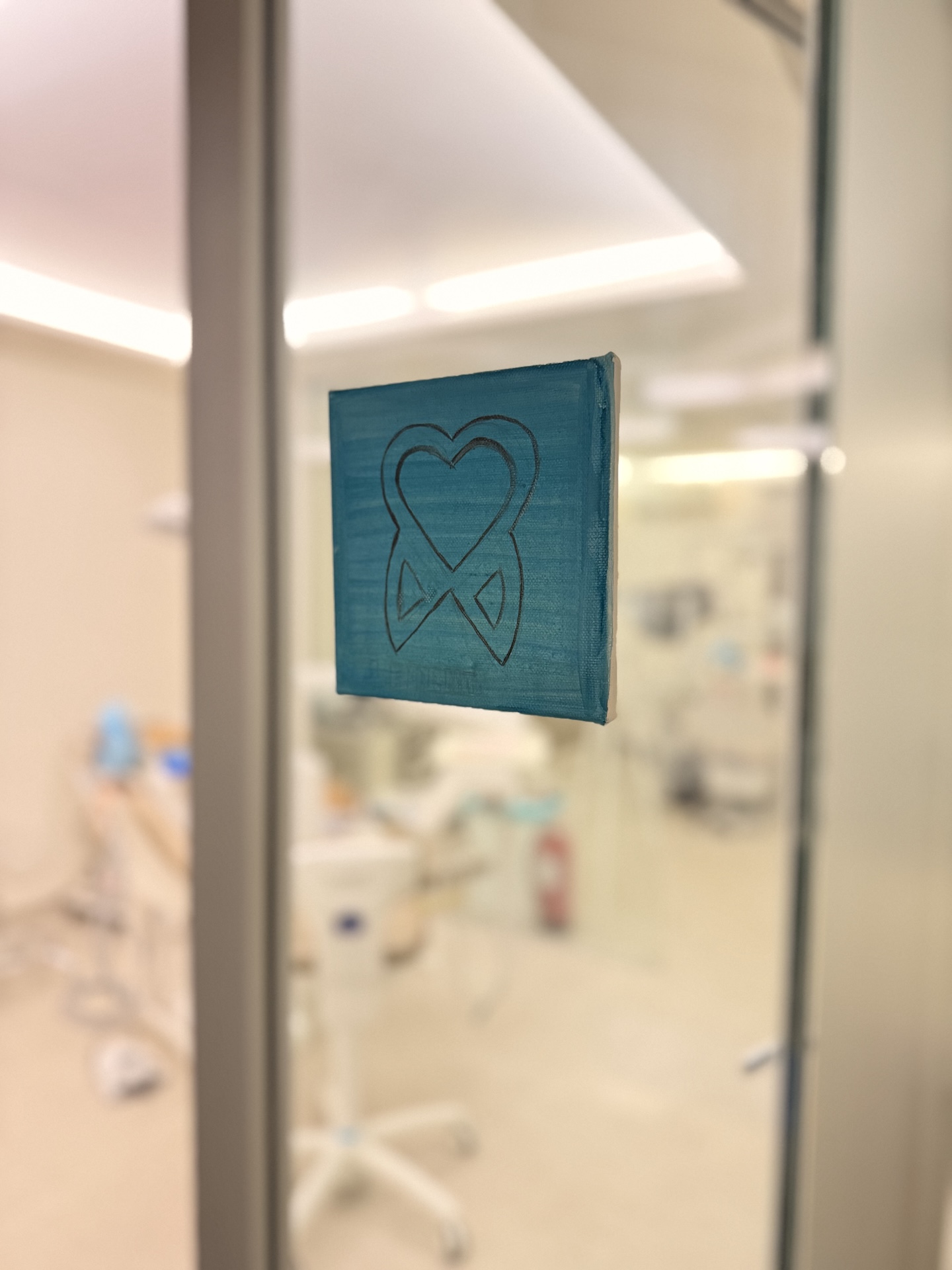 Blue square sign with a black outline of a tooth and a heart inside, mounted on a glass door of a dental office.