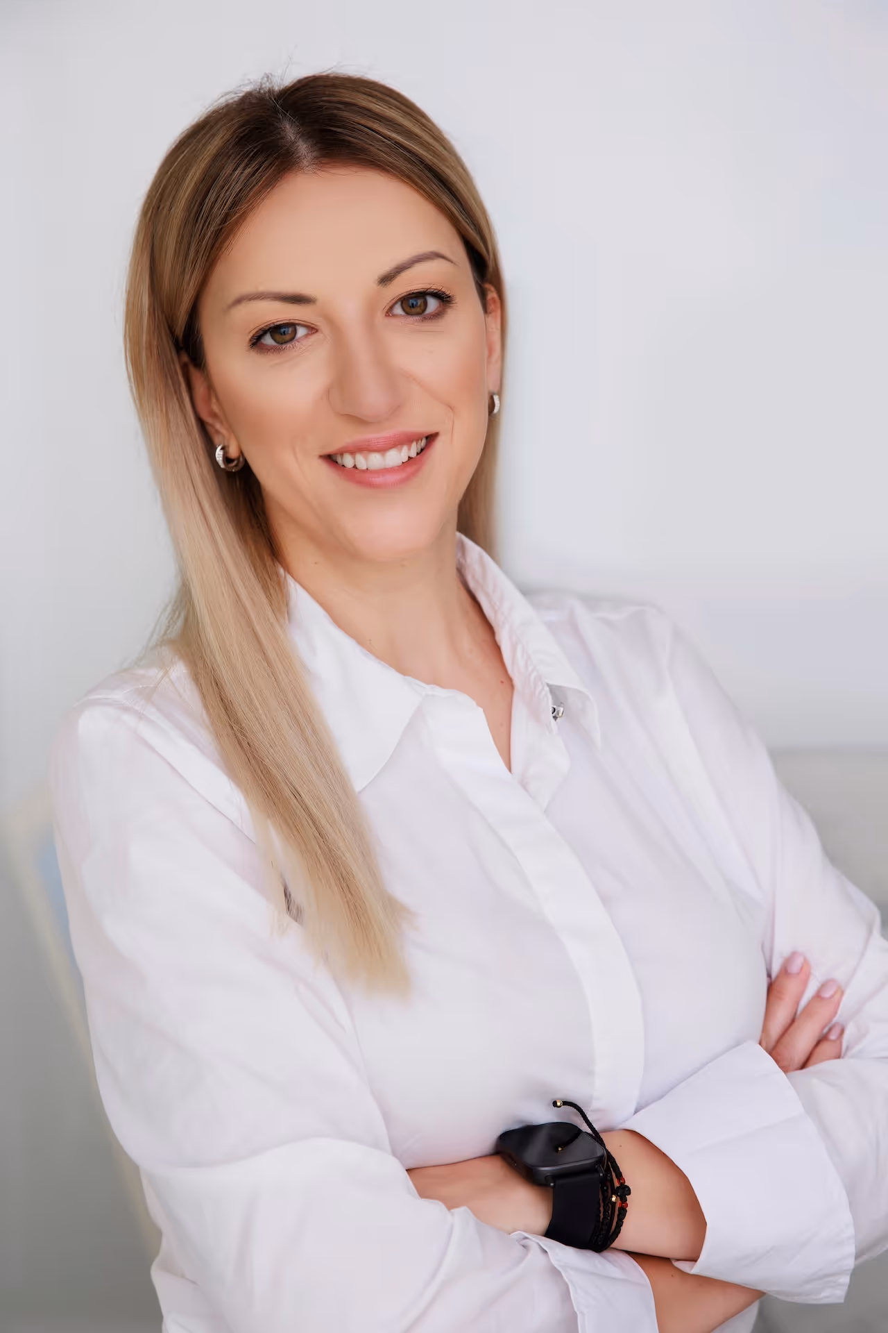 Smiling woman with straight blonde hair wearing a white shirt and a black smartwatch, arms crossed.