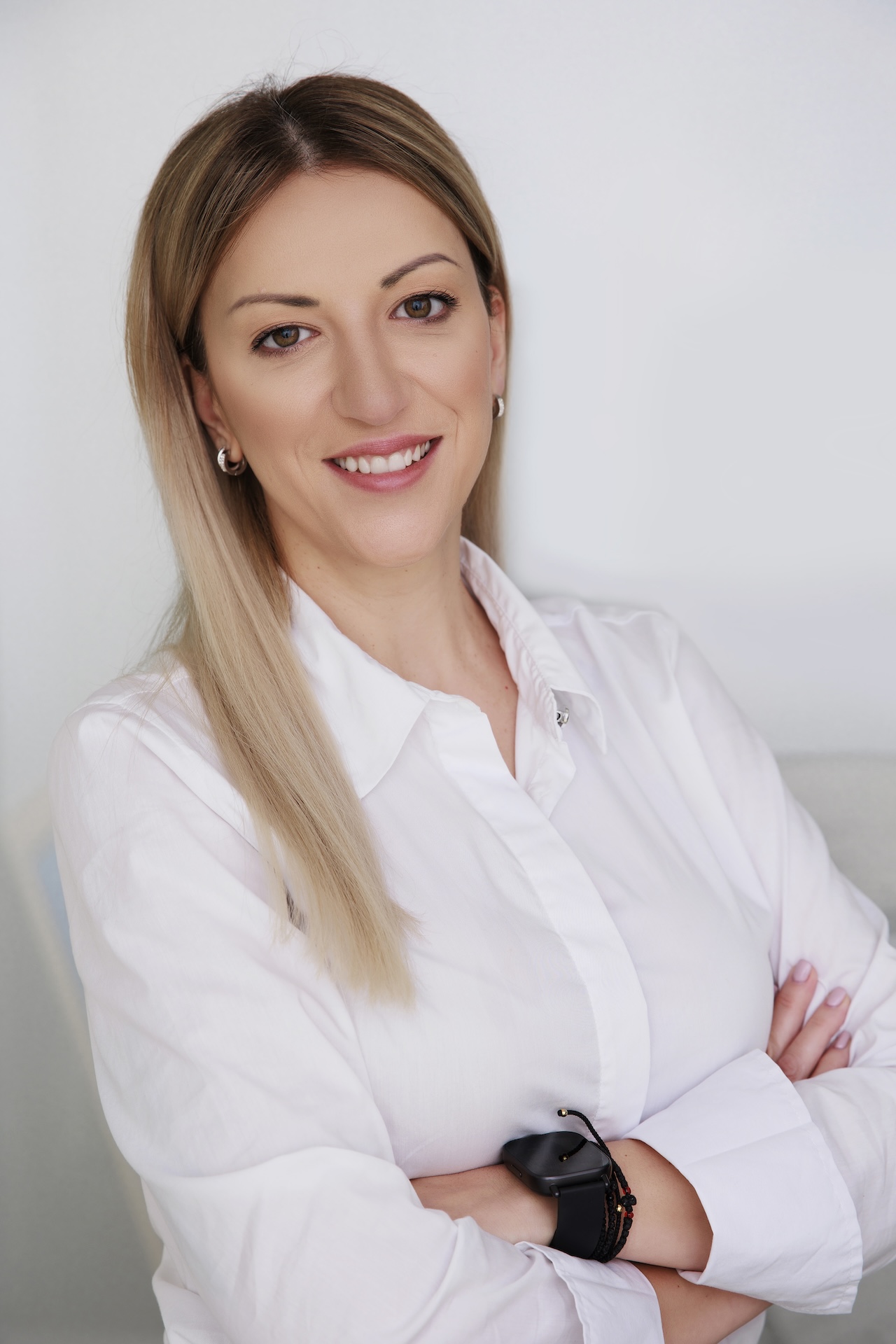Smiling woman with straight blonde hair wearing a white shirt and a black smartwatch, arms crossed.