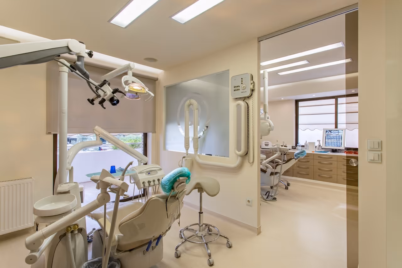 Modern dental clinic room with dentist chair, dental equipment, and stools near large windows with blinds.
