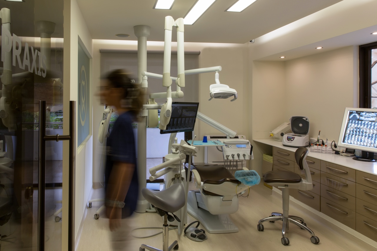 Modern dental clinic room with dental chair, equipment, monitors displaying X-rays, and a blurred person walking by glass door.