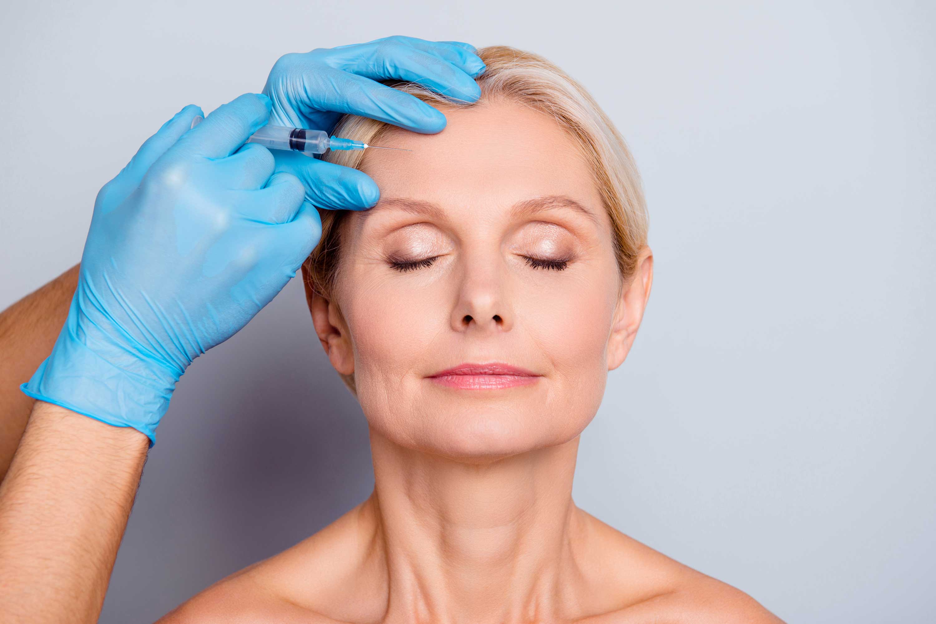 Close-up of a middle-aged woman with closed eyes receiving a cosmetic injection in her forehead from a person wearing blue gloves.