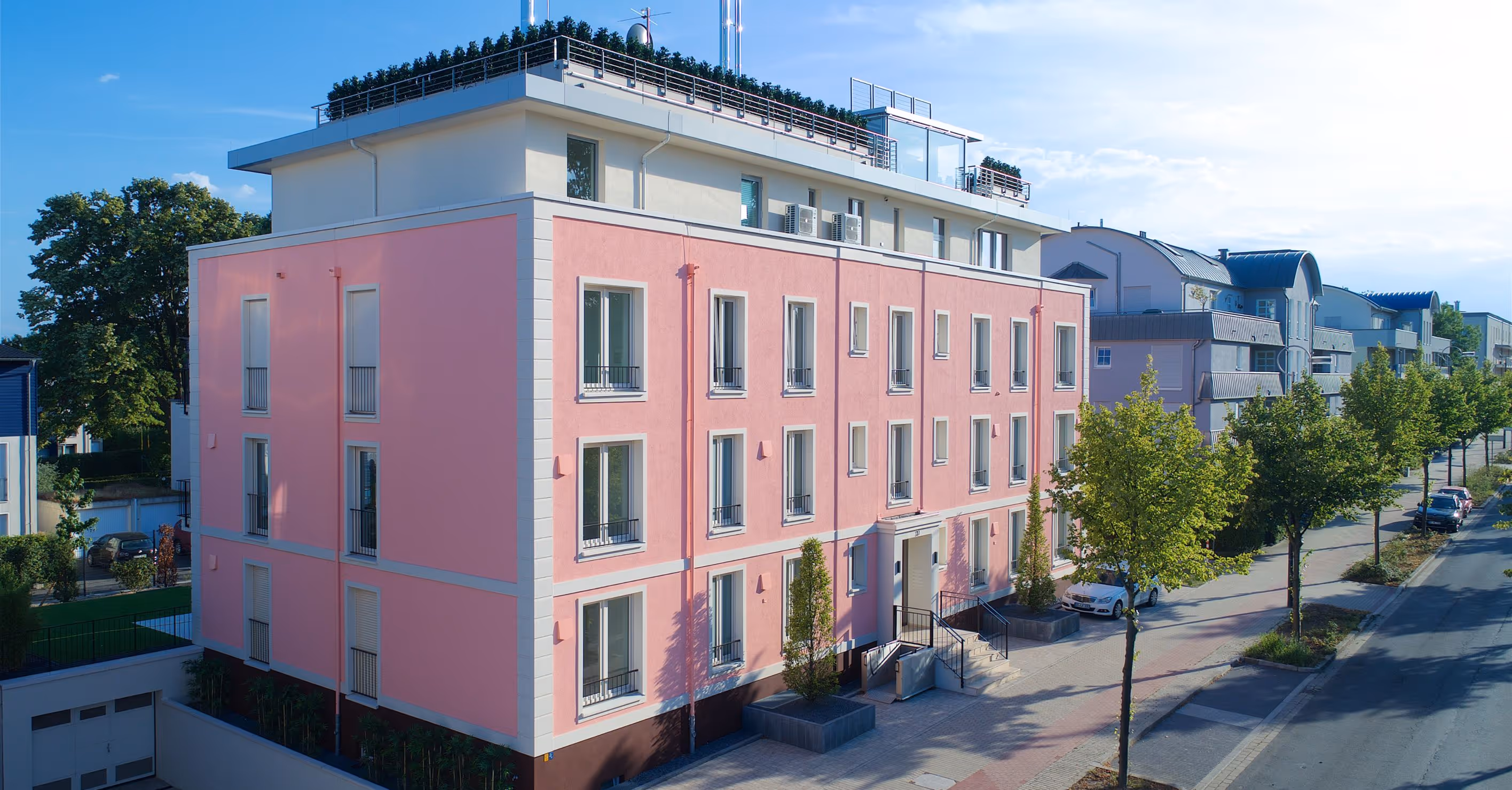 Ein modernes vierstöckiges Wohngebäude mit rosa Fassade an einer baumbestandenen Straße unter blauem Himmel.