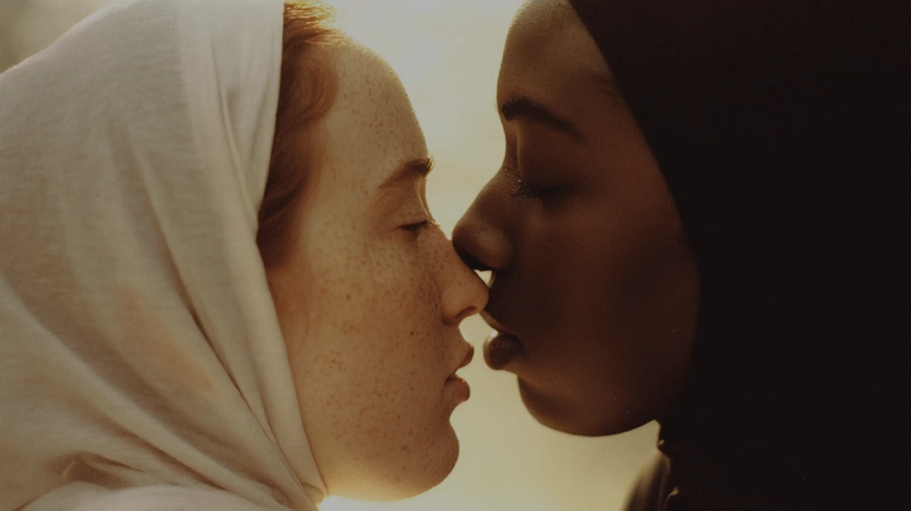 Zwei Personen mit Kopftüchern stehen eng beisammen, die Augen geschlossen, während eine zärtlich die Nase der anderen berührt.