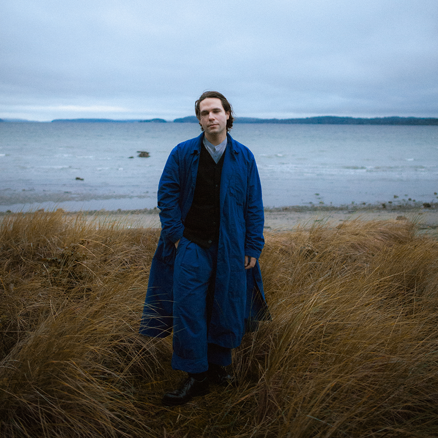 man in a blue blue overcoat and trousers walking in an overgrown field of tall yellow grass with ocean in background