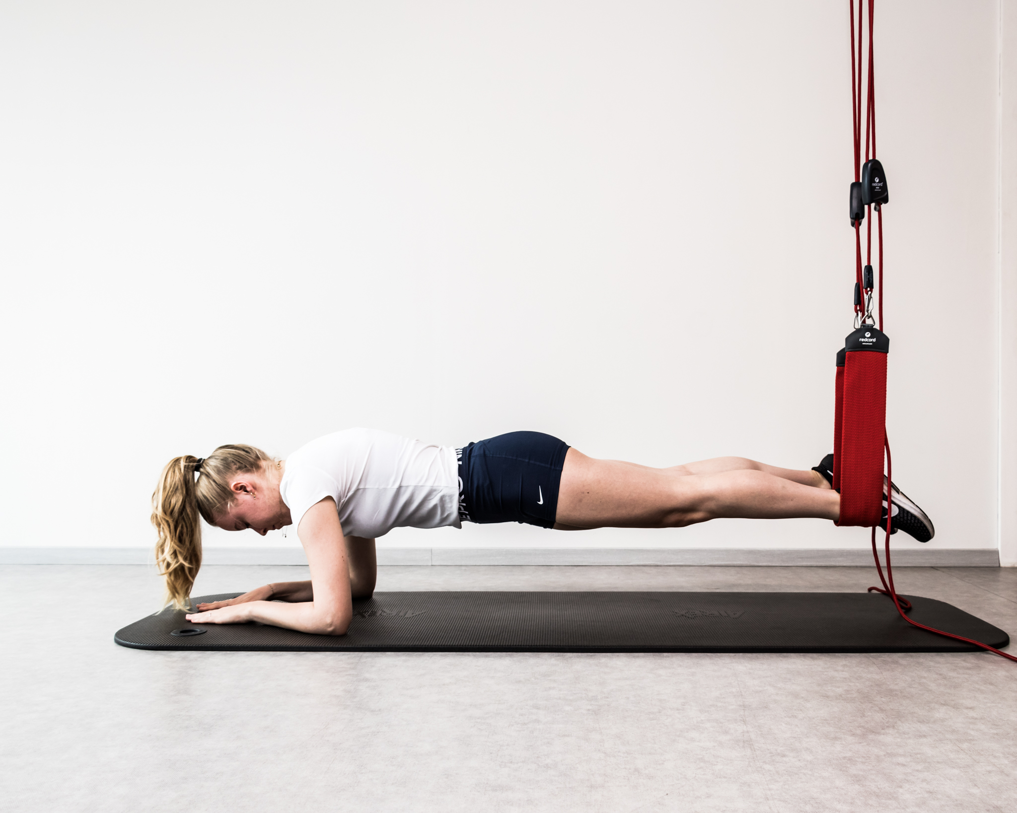 Vrouw voert plank-oefening uit met haar voeten in rode fitnessbanden aan de muur, liggend op een zwarte yogamat.