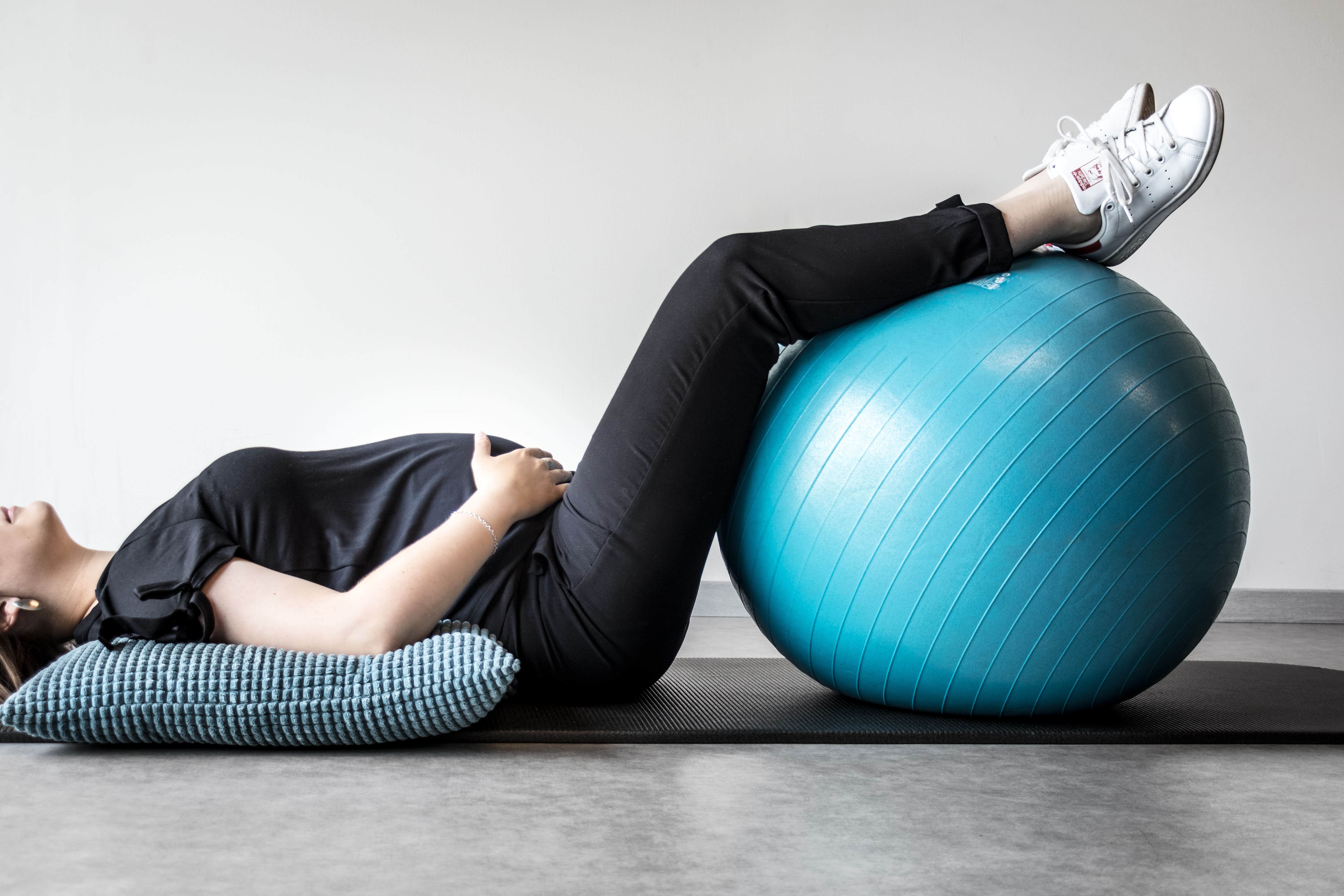 Zwangere vrouw ligt op een mat met haar benen op een blauwe fitnessbal en haar hoofd op een kussen.