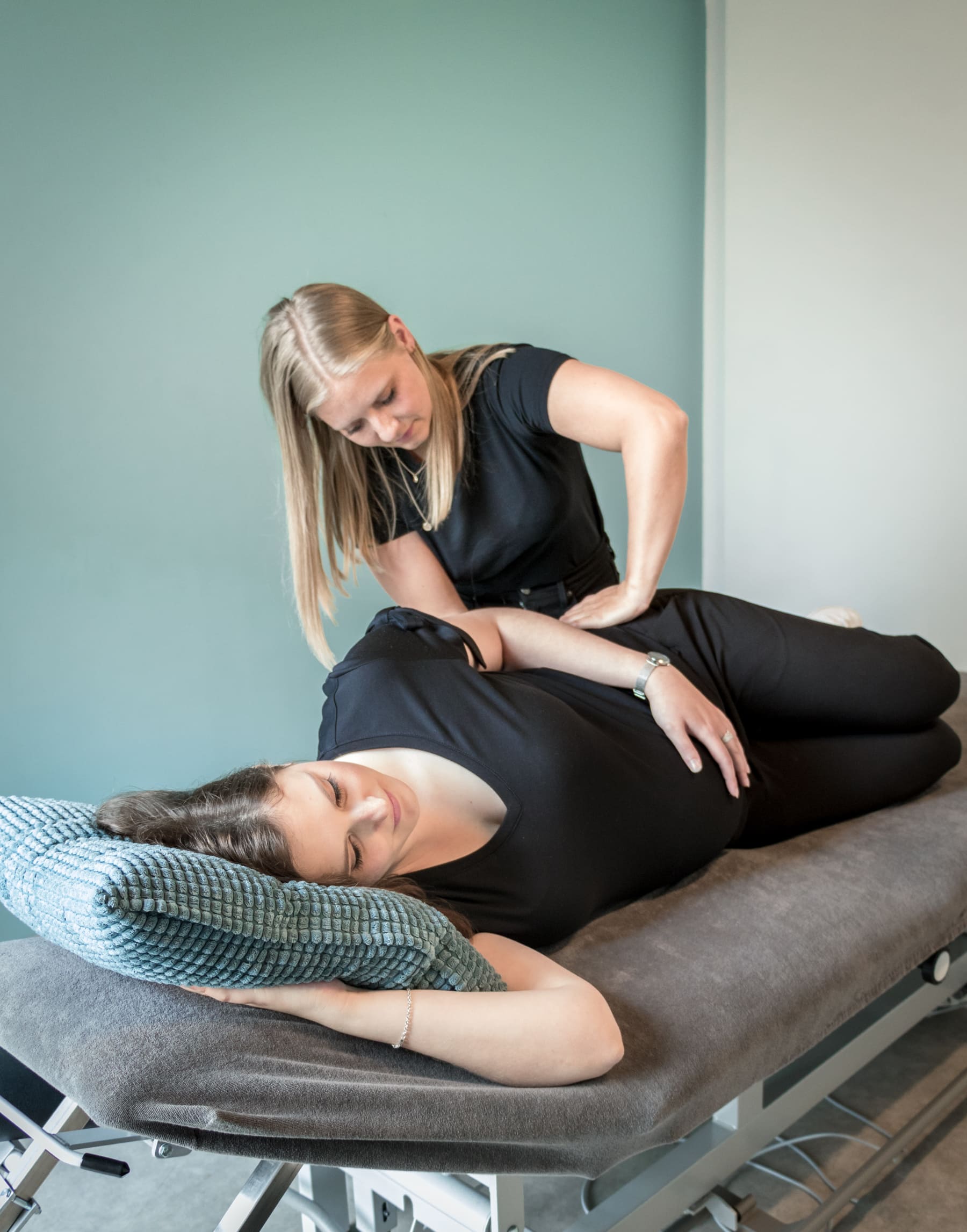 Een vrouw ligt op haar zij op een behandelbank met een kussen onder haar hoofd terwijl een andere vrouw haar rug behandeld.