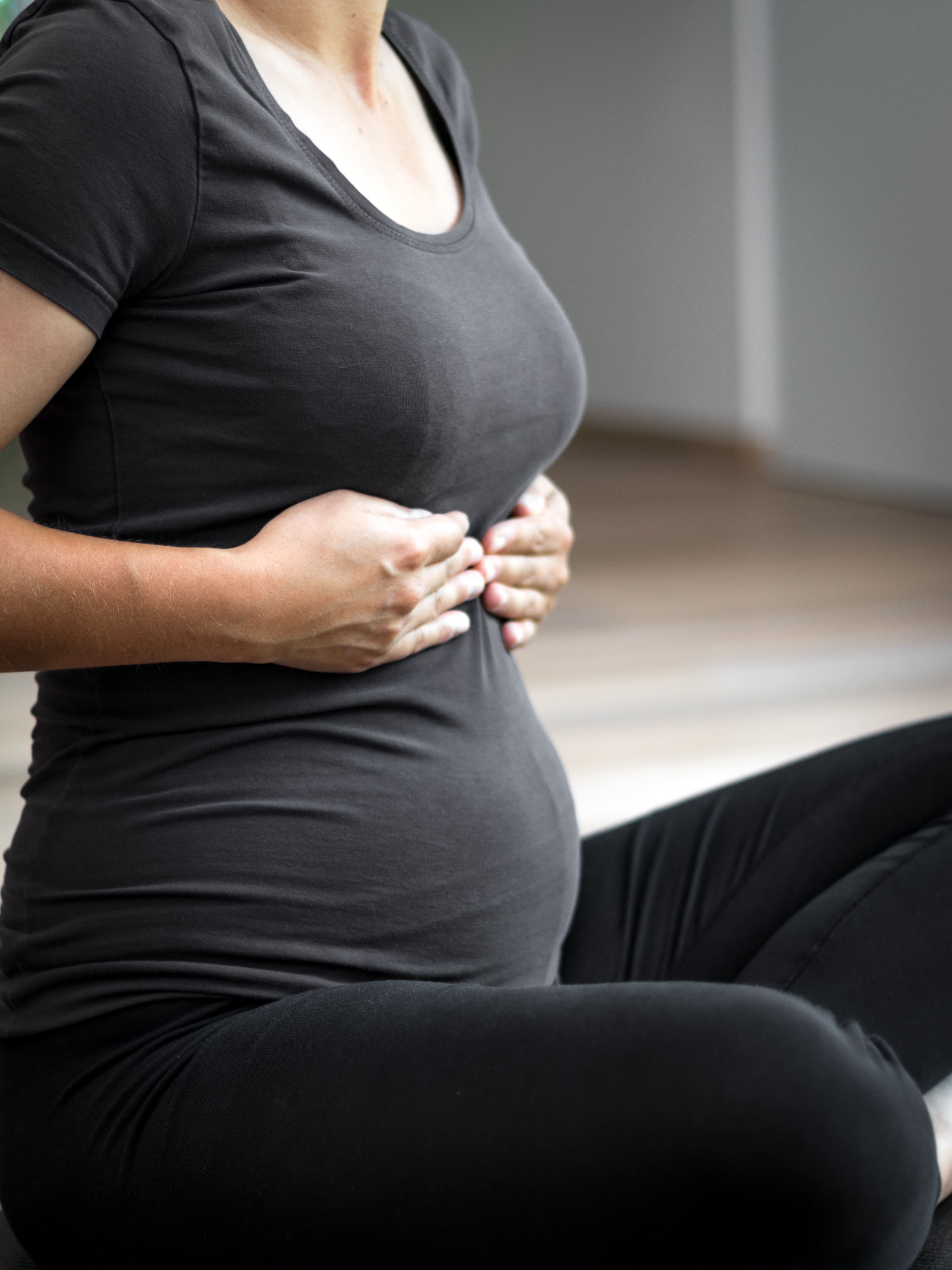 Zwangere vrouw zittend met handen op haar borst en buik, gekleed in een zwart shirt en zwarte broek.