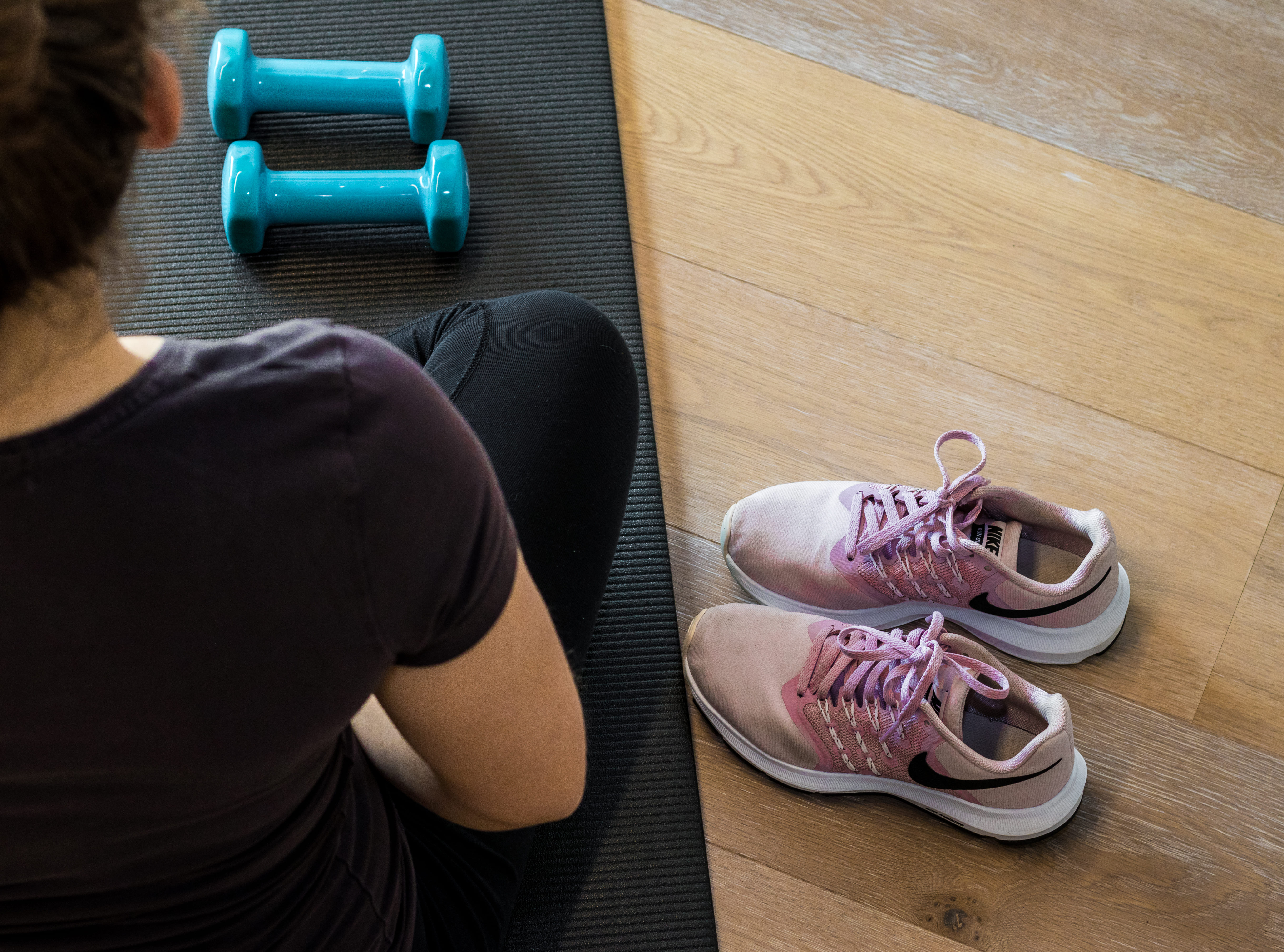 Vrouw zittend op een zwarte fitnessmat met twee blauwe dumbbells en een paar roze Nike sportschoenen op de houten vloer.