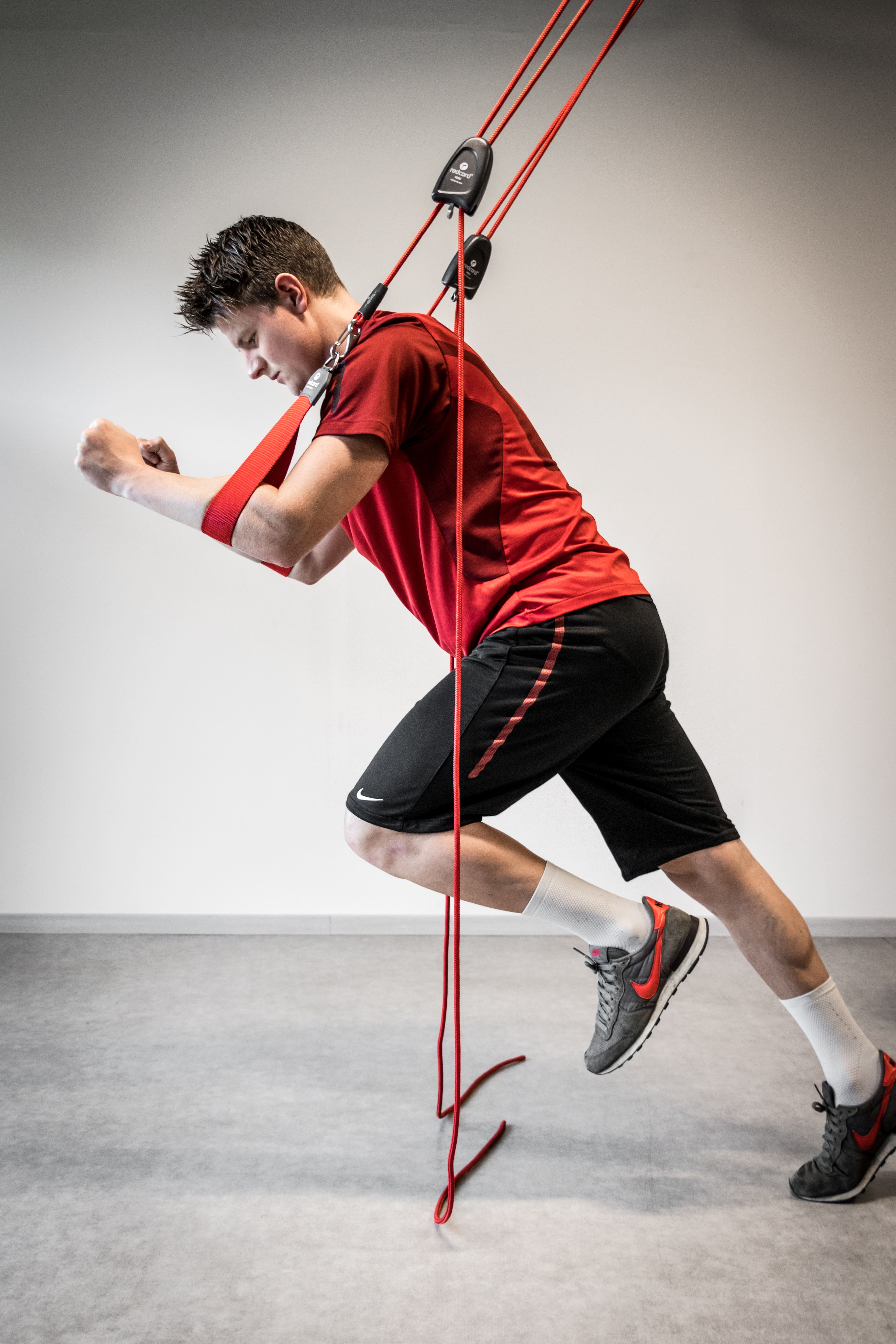 Jongen in rood sportshirt en zwarte shorts traint met een rood elastisch touw dat aan een harnas vastzit, alsof hij vooruit rent.