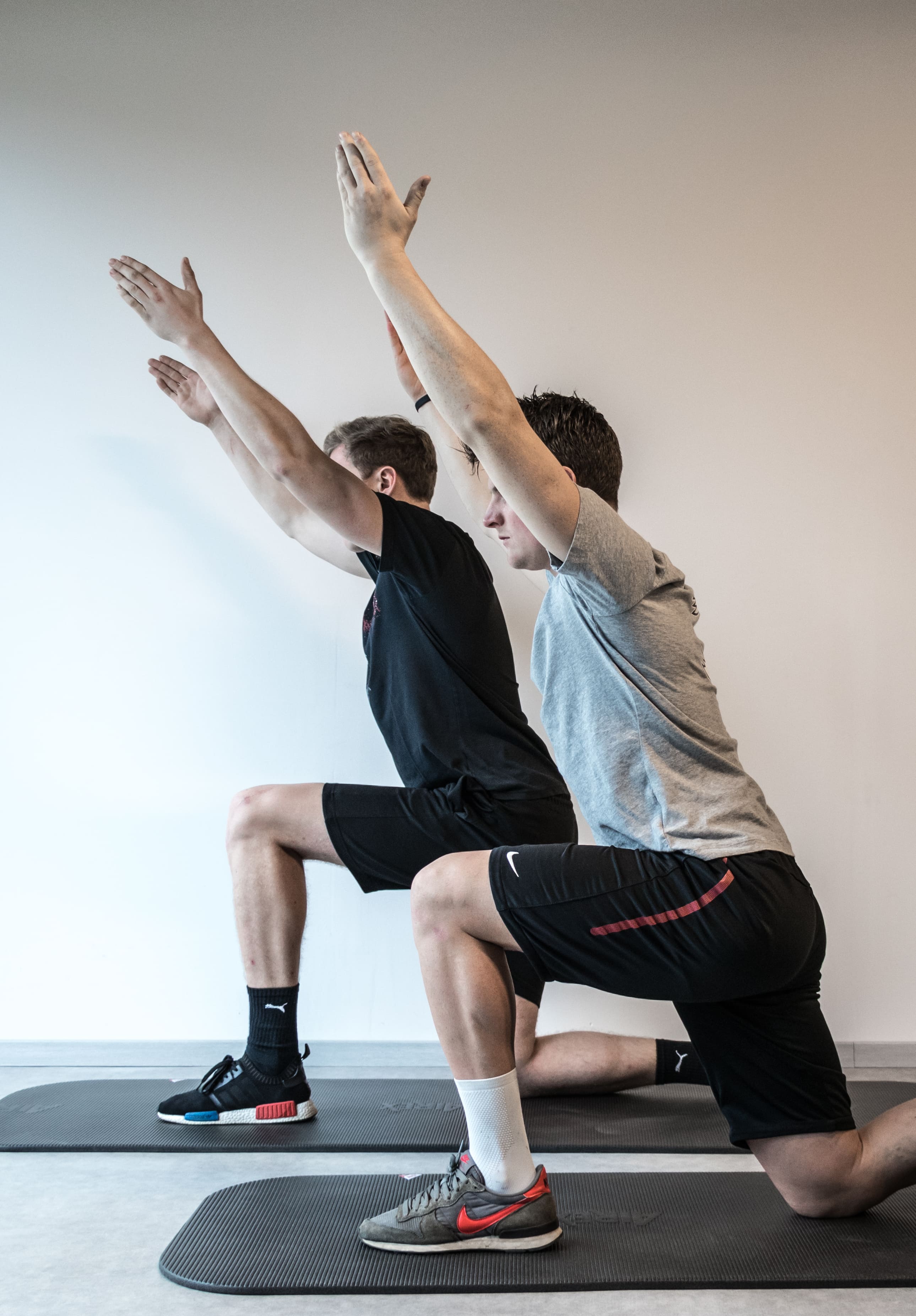 Twee jonge mannen doen lunges met opgestrekte armen op yogamatten in een lichte kamer.