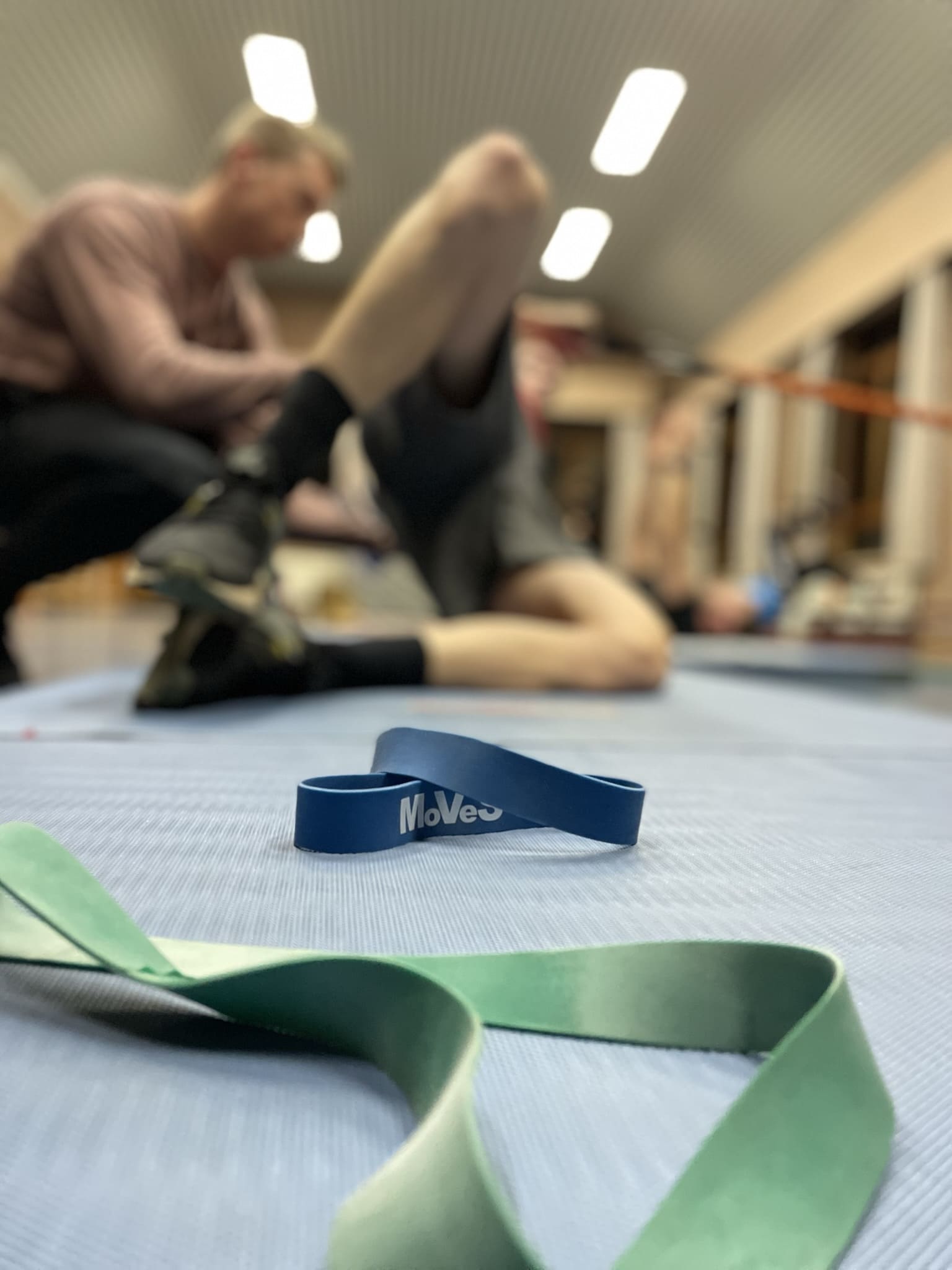 Een gymzaal met weerstandsbanden op de mat en personen die stretchen en trainen op de achtergrond.