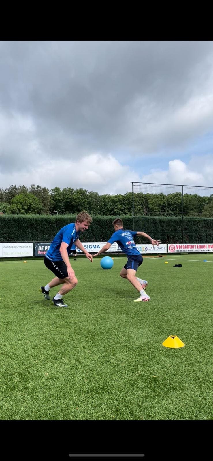Twee jongens in blauwe sportkledij trainen op een voetbalveld met groene grasmat onder een bewolkte hemel.