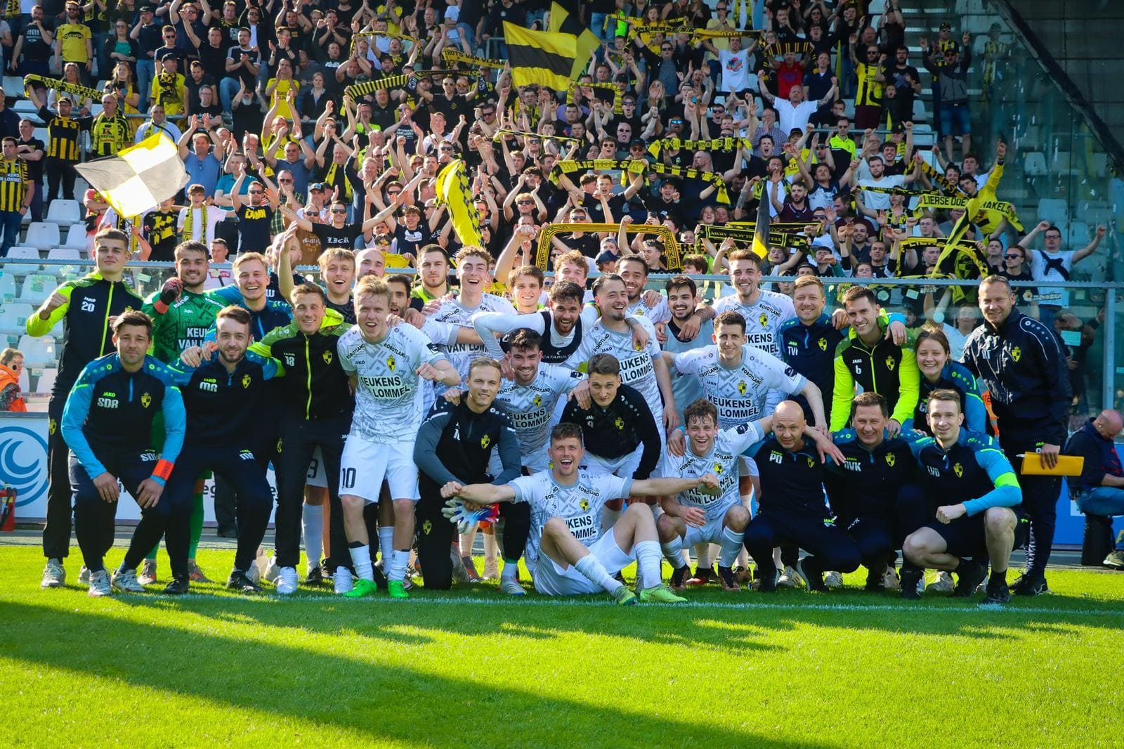 Jubelend voetbalteam in witte tenues op het veld met enthousiaste supporters op de achtergrond die gele en zwarte sjaals en vlaggen zwaaien.