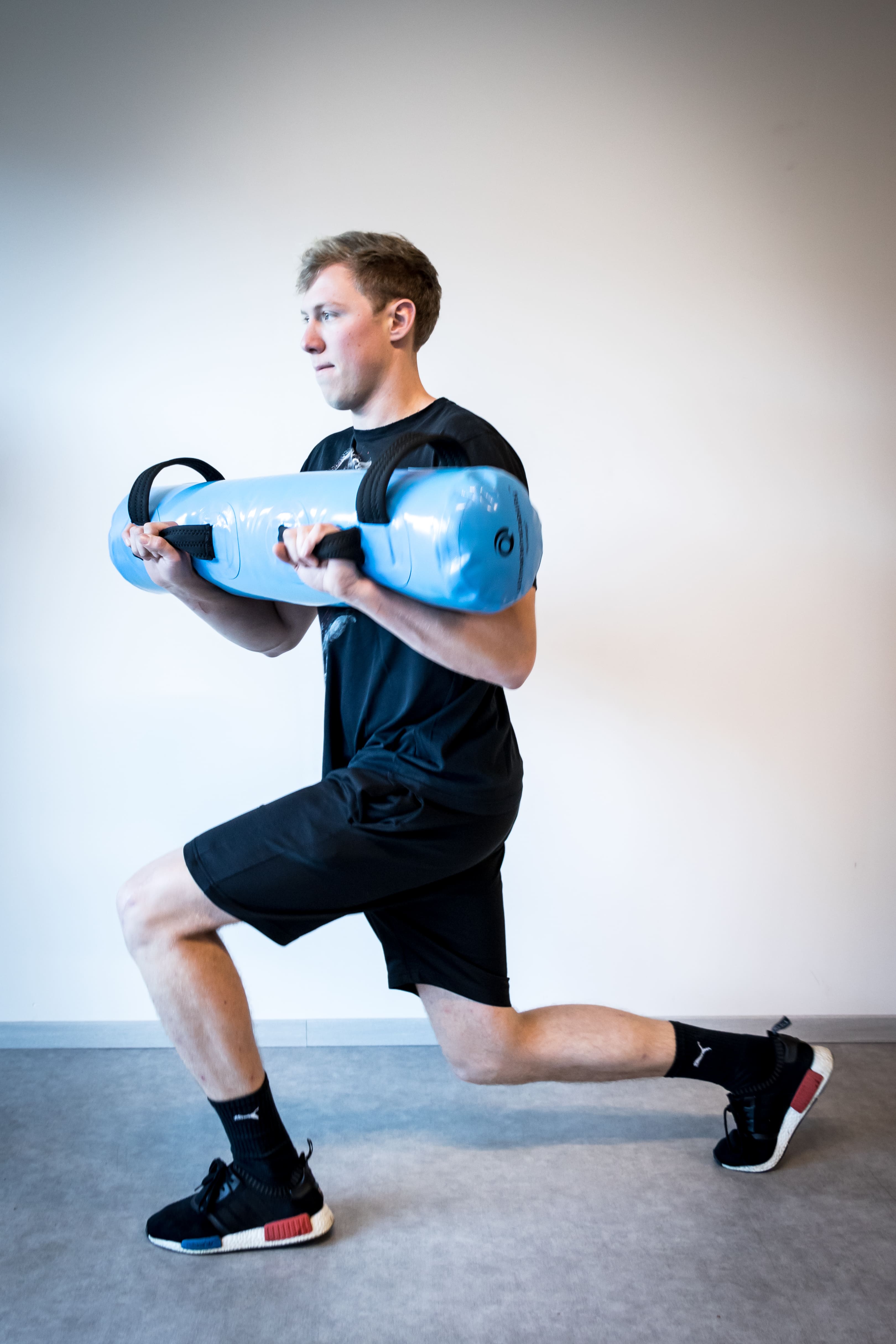Een man voert een wandelende lunge uit met een blauwe sandbag in een lichte kamer.