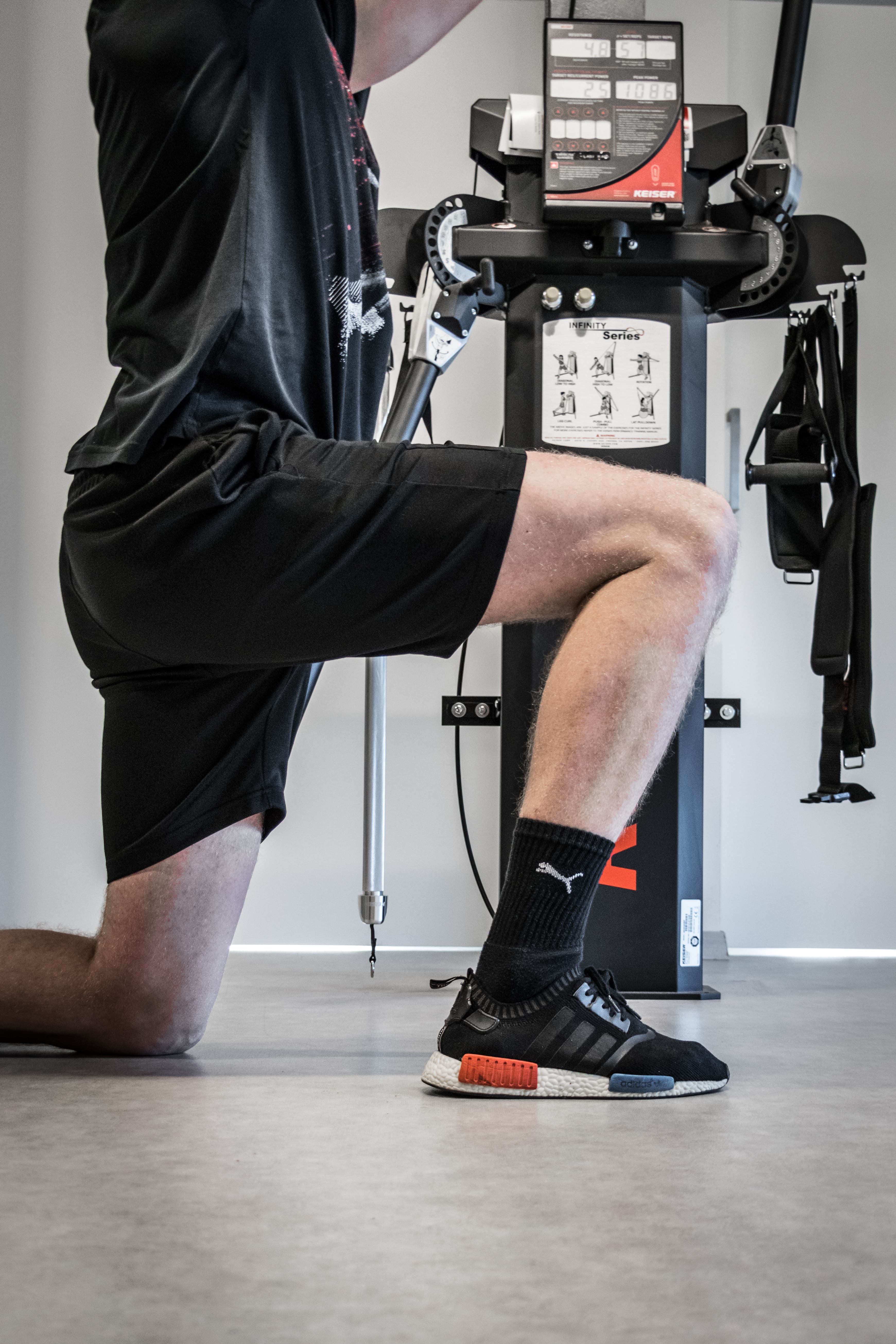 Person knielt op één knie tijdens het sporten in een fitnessruimte met krachttrainingsapparaat op de achtergrond.