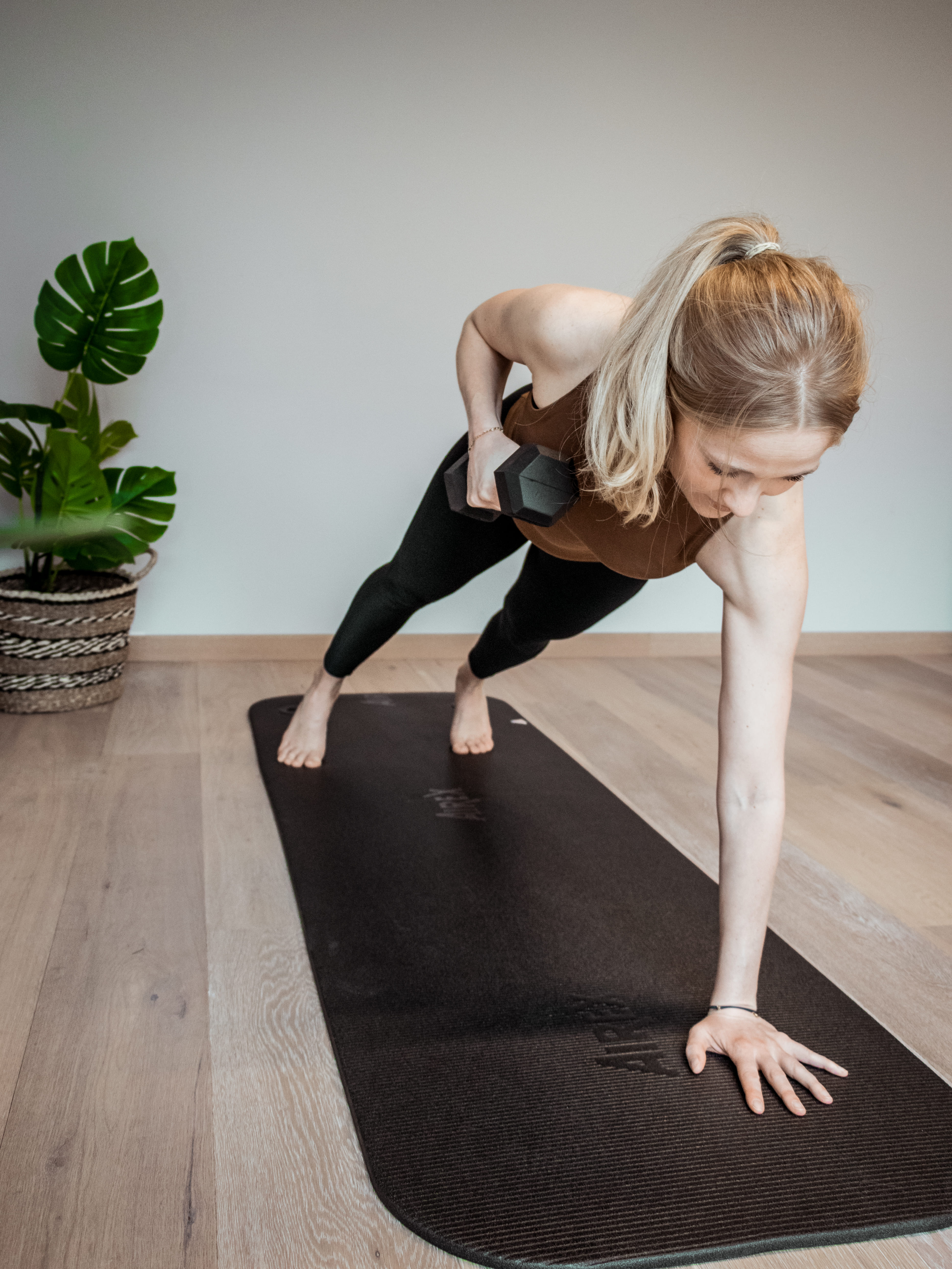 Vrouw in sportkleding doet plank met één hand op een yogamat terwijl ze in de andere hand een halter vasthoudt.