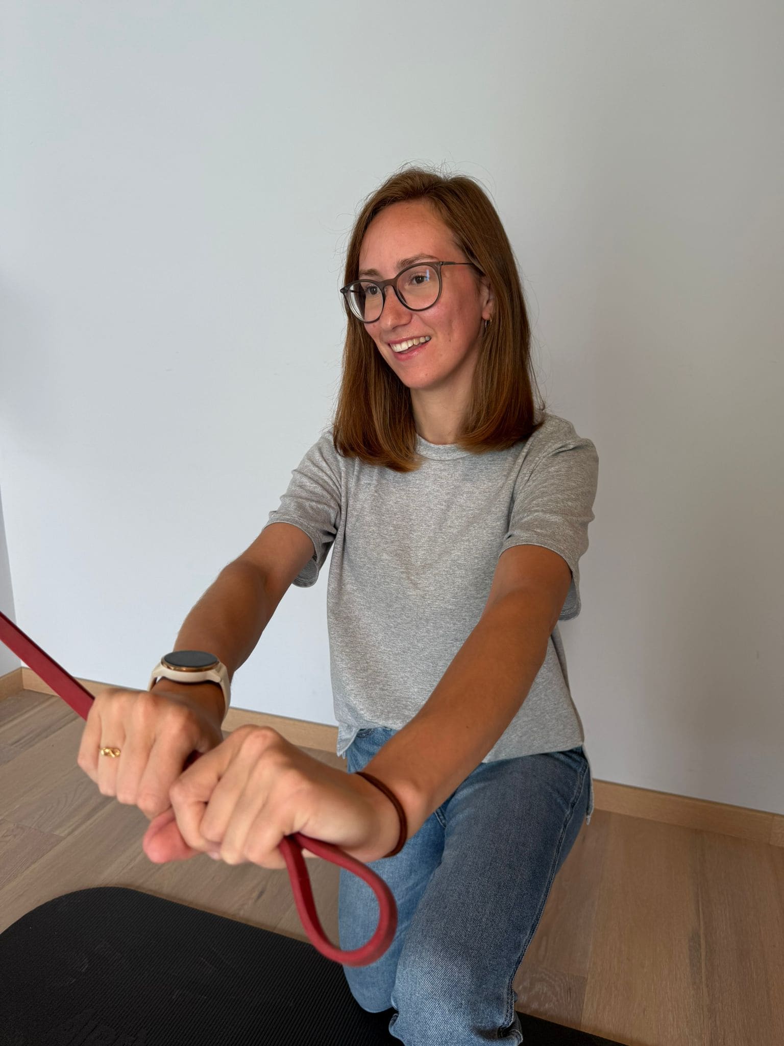 Vrouw met bril en grijs T-shirt die knielt en een rode oefenband vasthoudt in een kamer met houten vloer.