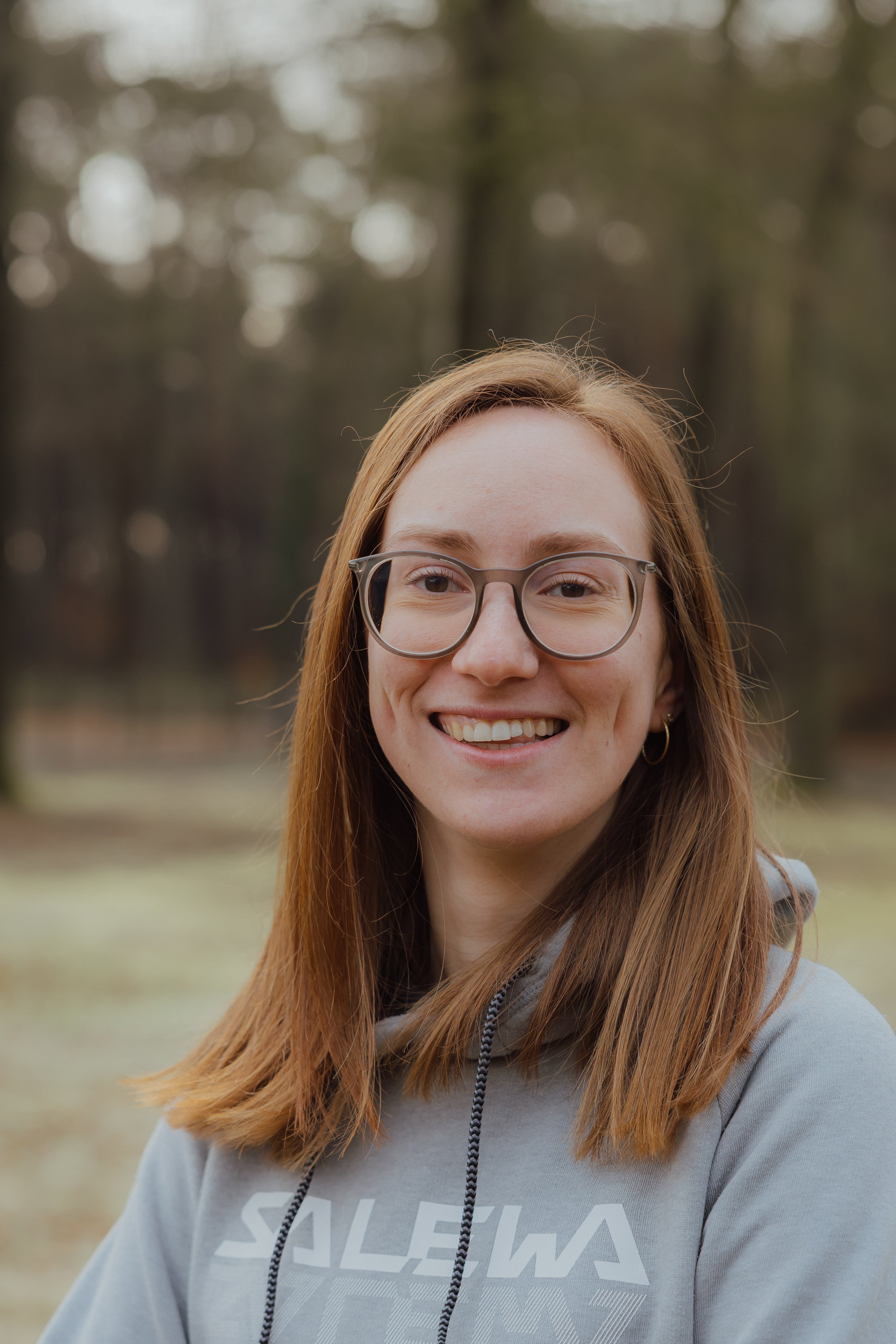Vrouw met lang rood haar, bril en grijze Salewa hoodie glimlachend buiten in een bosrijke omgeving.