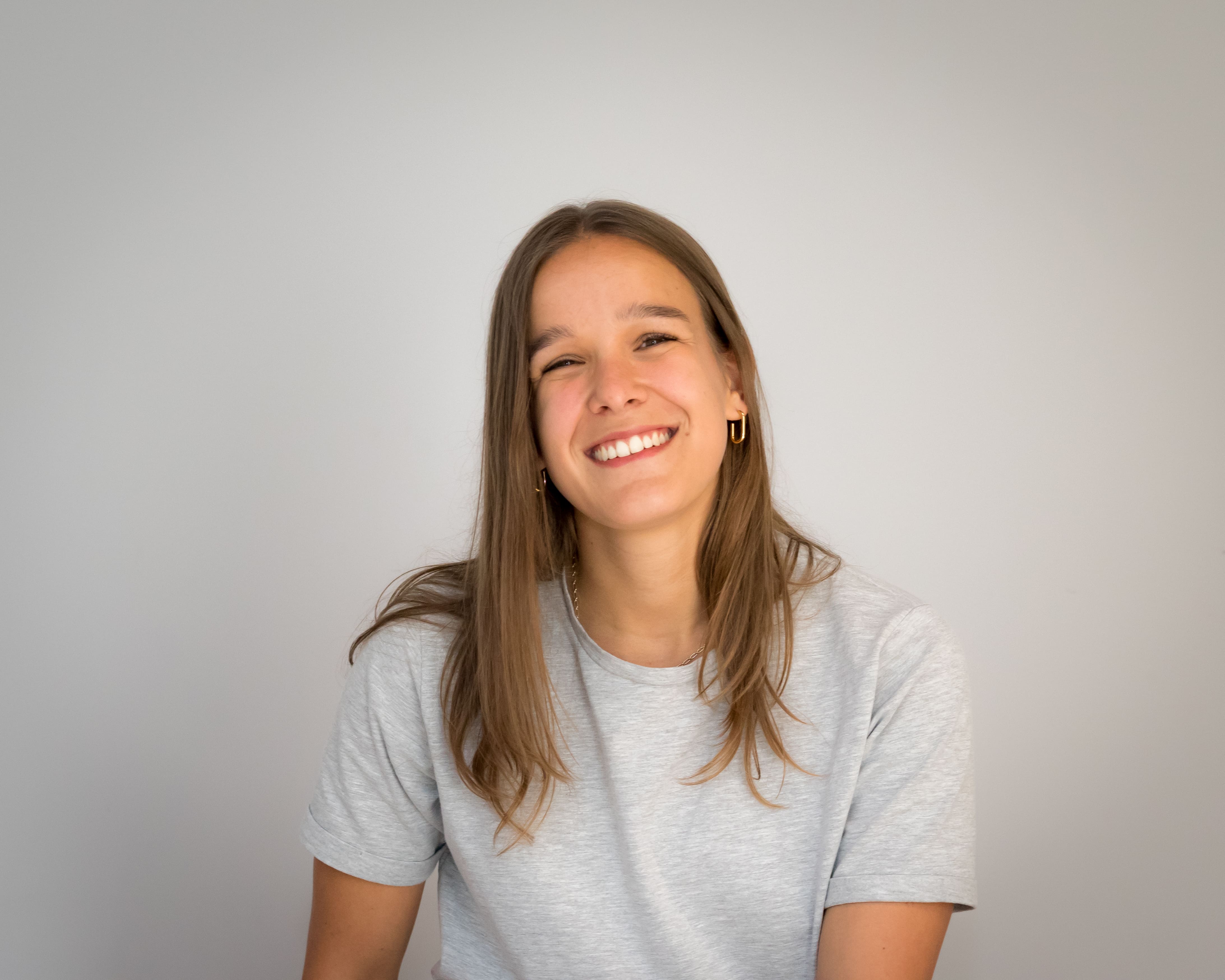 Jonge vrouw met lang bruin haar lacht naar de camera tegen een lichte achtergrond.