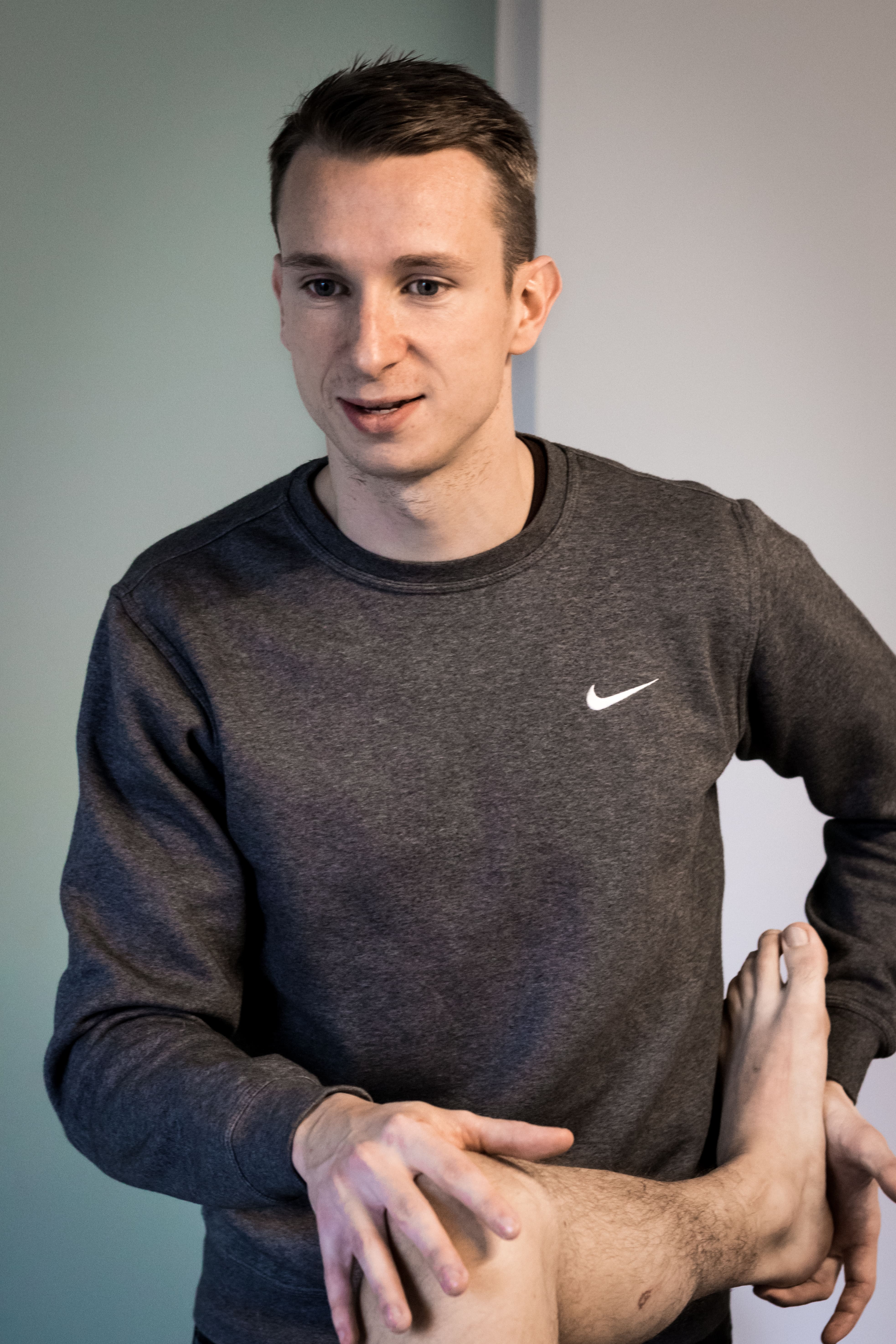 Man in een grijze Nike-trui die het been van een andere persoon vasthoudt tijdens een fysiotherapie-sessie.