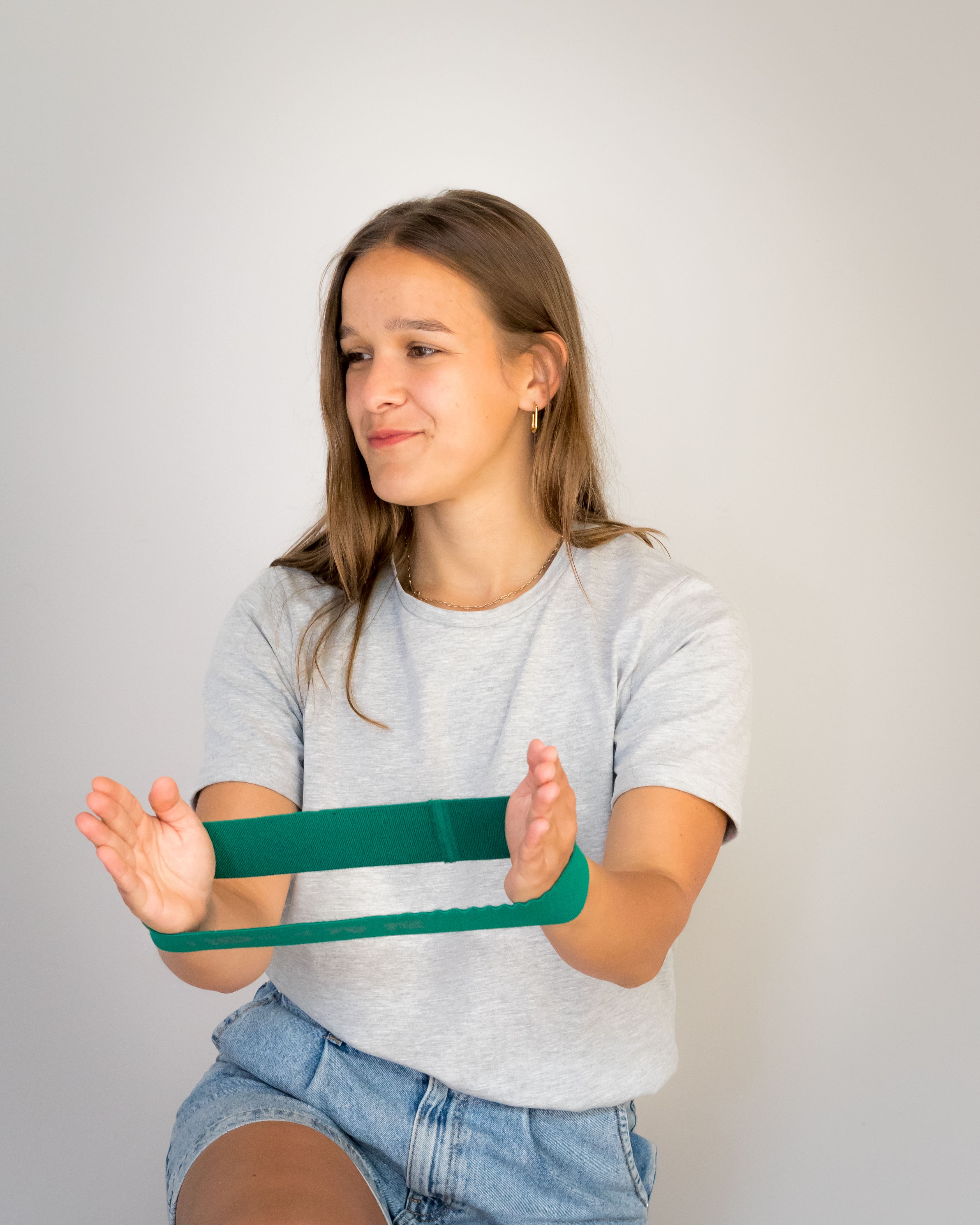 Jonge vrouw in grijs T-shirt en jeans short die een groene weerstandsband tussen haar handen uitrekt.