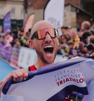 Jubelende triatleet met zonnebril die een finishlint vasthoudt tijdens een race, met toeschouwers op de achtergrond.