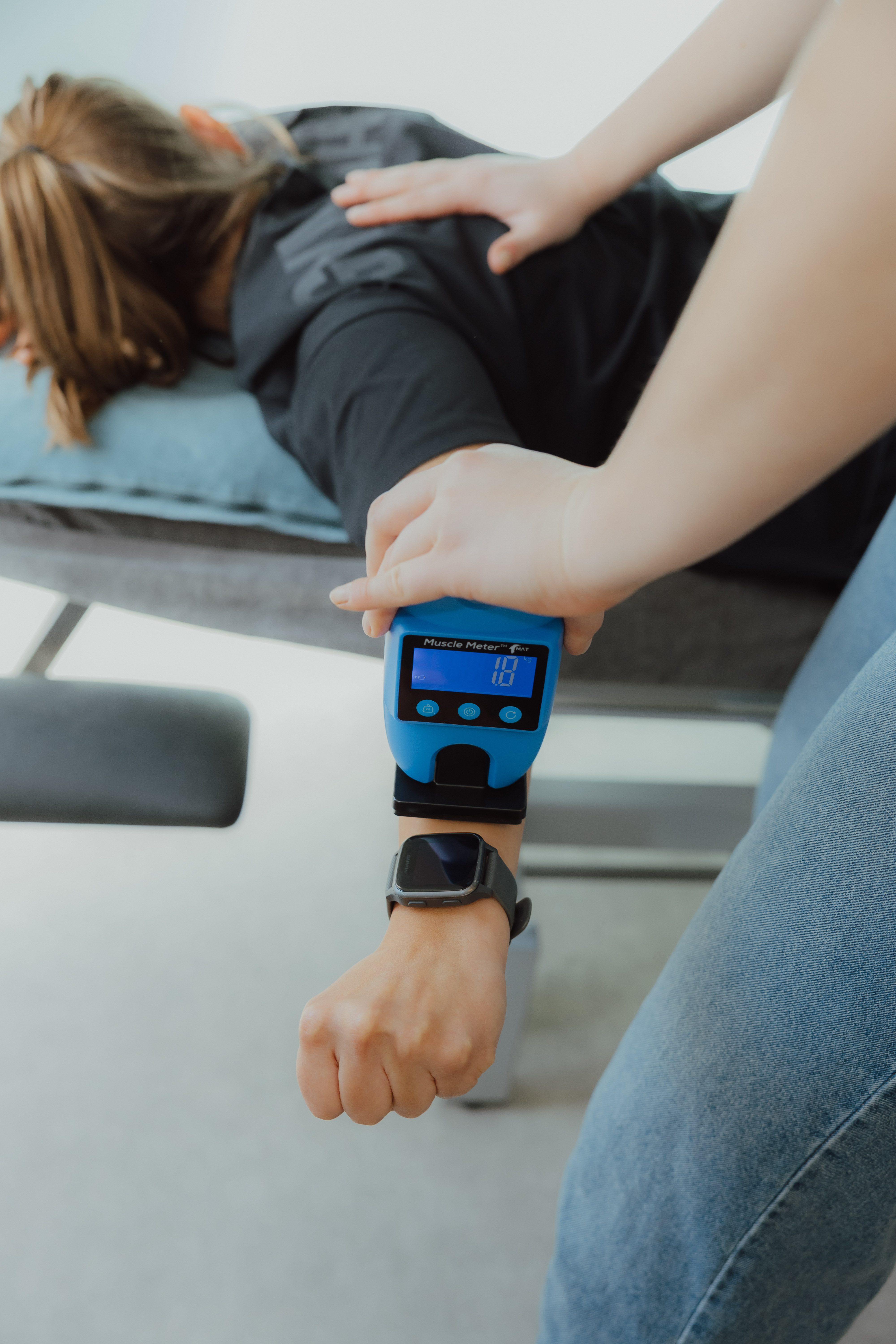 Vrouw ligt op tafel terwijl iemand een spierkrachtmeter op haar pols gebruikt, scherm toont 18 kg.