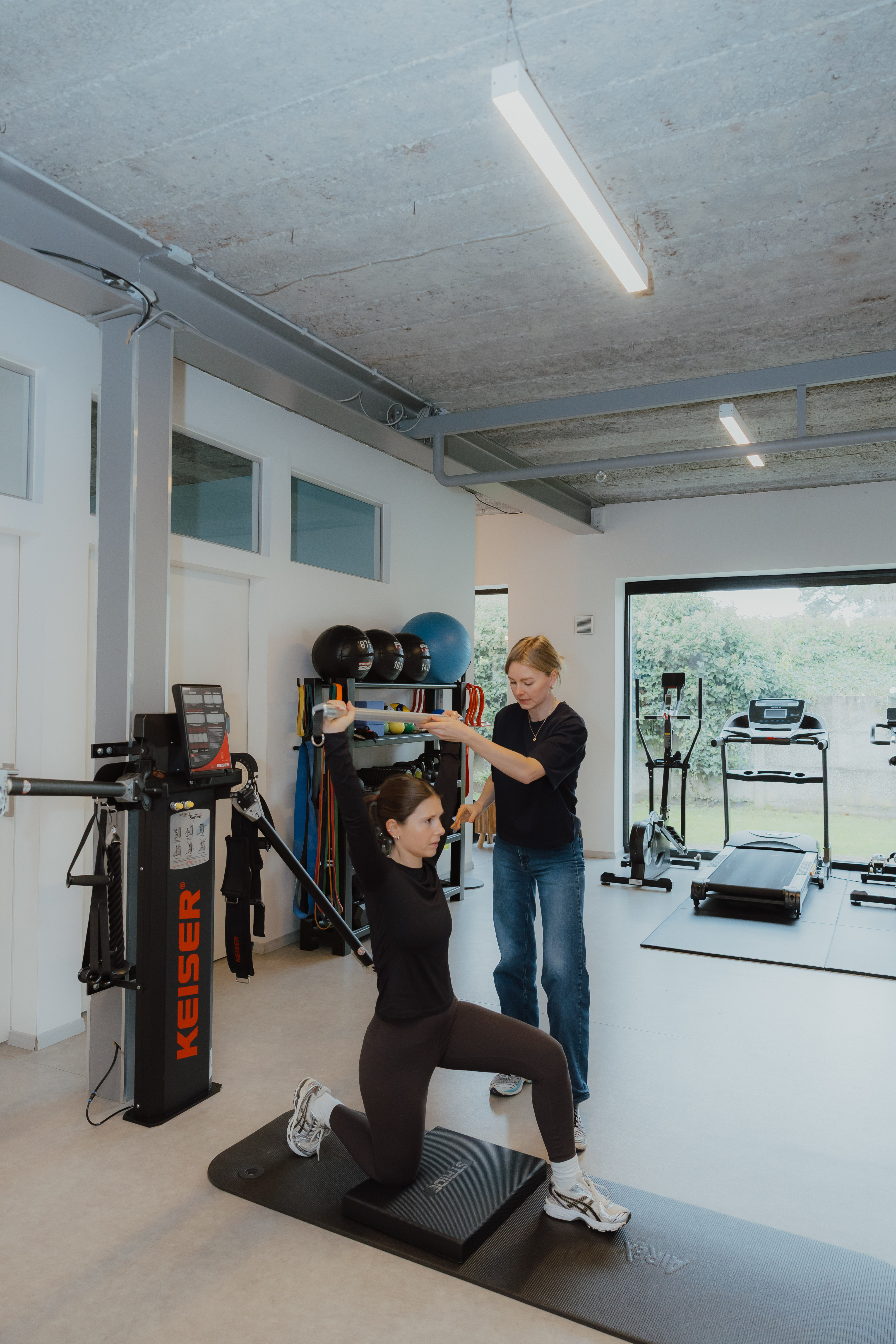 Fysiotherapeut begeleidt een vrouw die op een mat knielt en een gewicht boven haar hoofd houdt in een goed uitgeruste oefenruimte.
