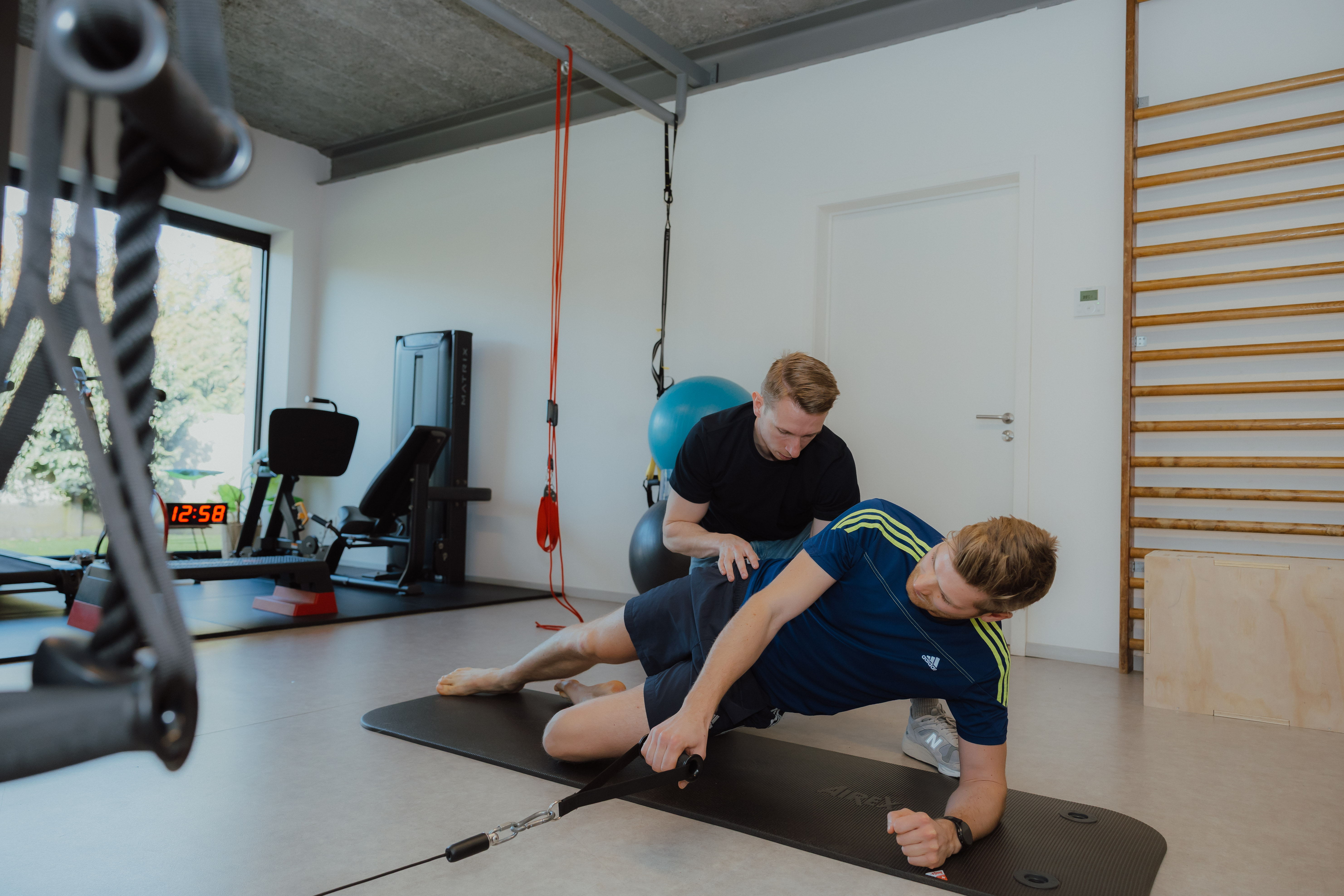 Man in sportkleding doet zijwaartse oefening met kabel, begeleid door een trainer in een fitnessruimte.